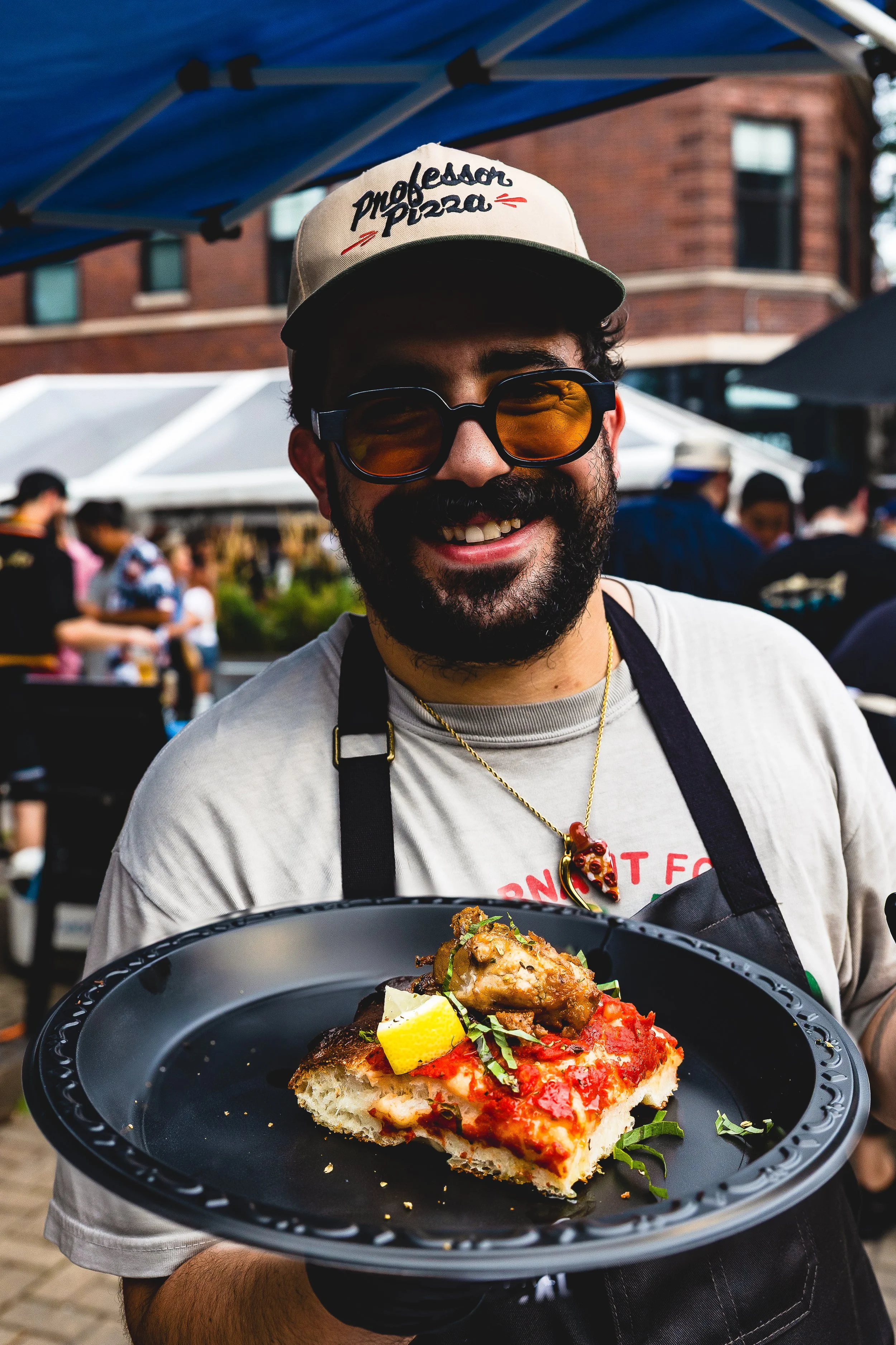 A man wearing a cap that reads 'Professor Pizza', sunglasses, and a necklace is holding a black plate with a slice of pizza topped with vegetables and chicken, smiling at an outdoor event with tents and people in the background.
