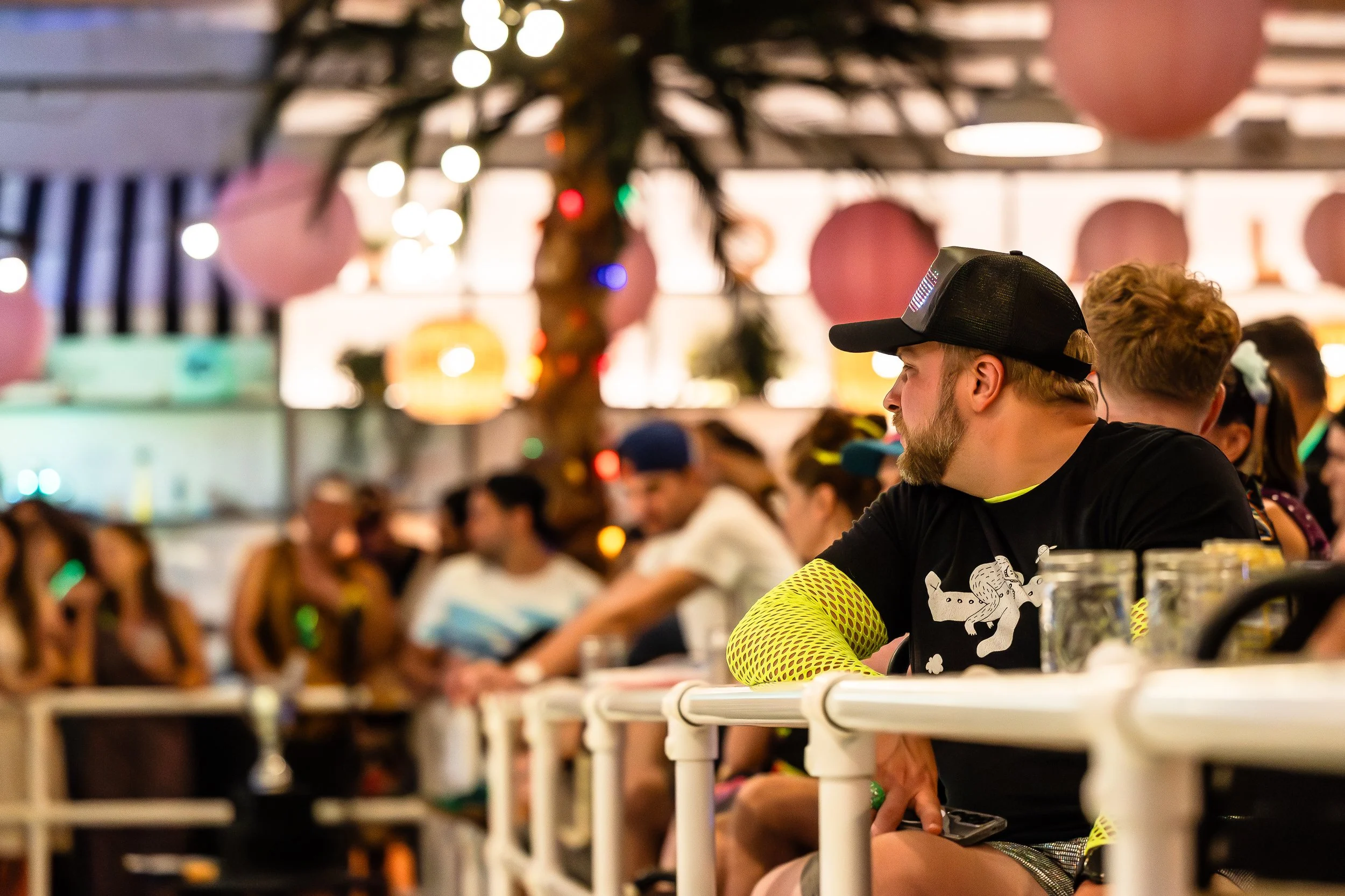 A man with a beard and a black cap sitting at a bar or counter, surrounded by other people in a lively indoor setting decorated with pink and paper lanterns.