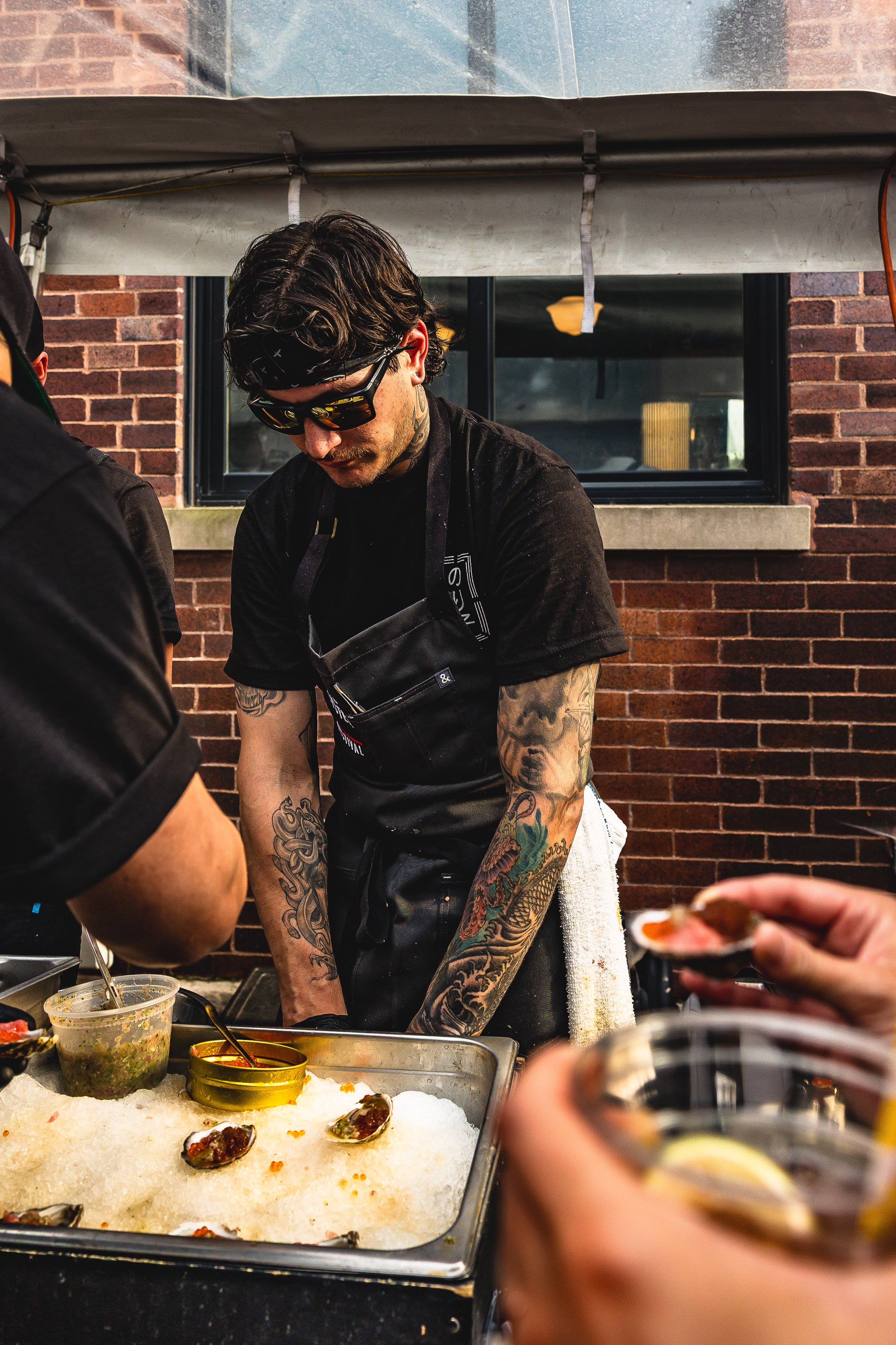 A tattooed man wearing sunglasses and a black apron preparing food at an outdoor event, with a brick building and window in the background.