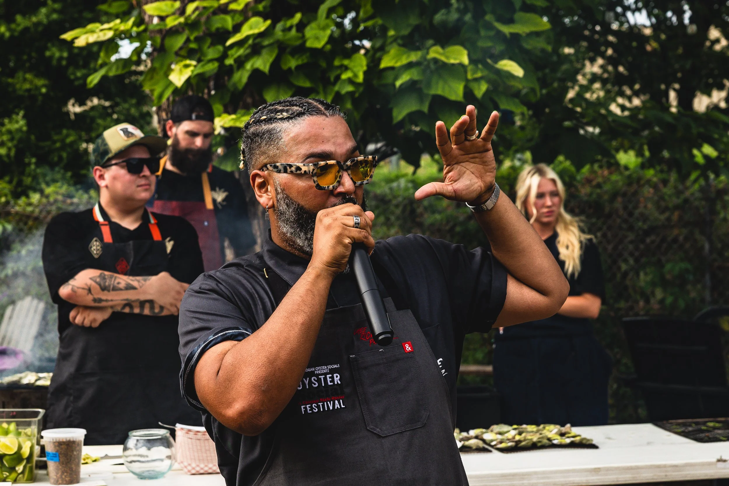A man with braided hair, glasses, and a beard holding a microphone and speaking at an outdoor event with a group of people behind him.