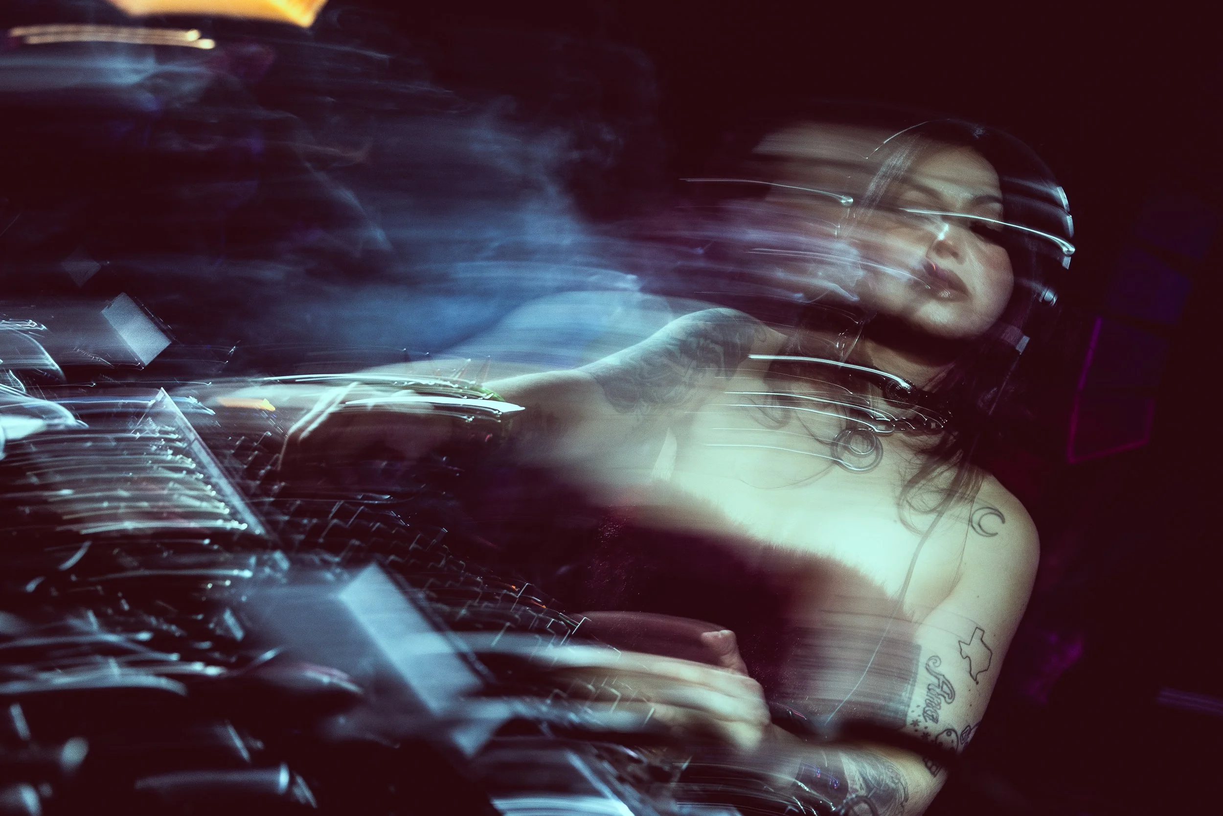 A woman with tattoos wearing headphones, operating DJ equipment in a dark environment with motion blur effects.