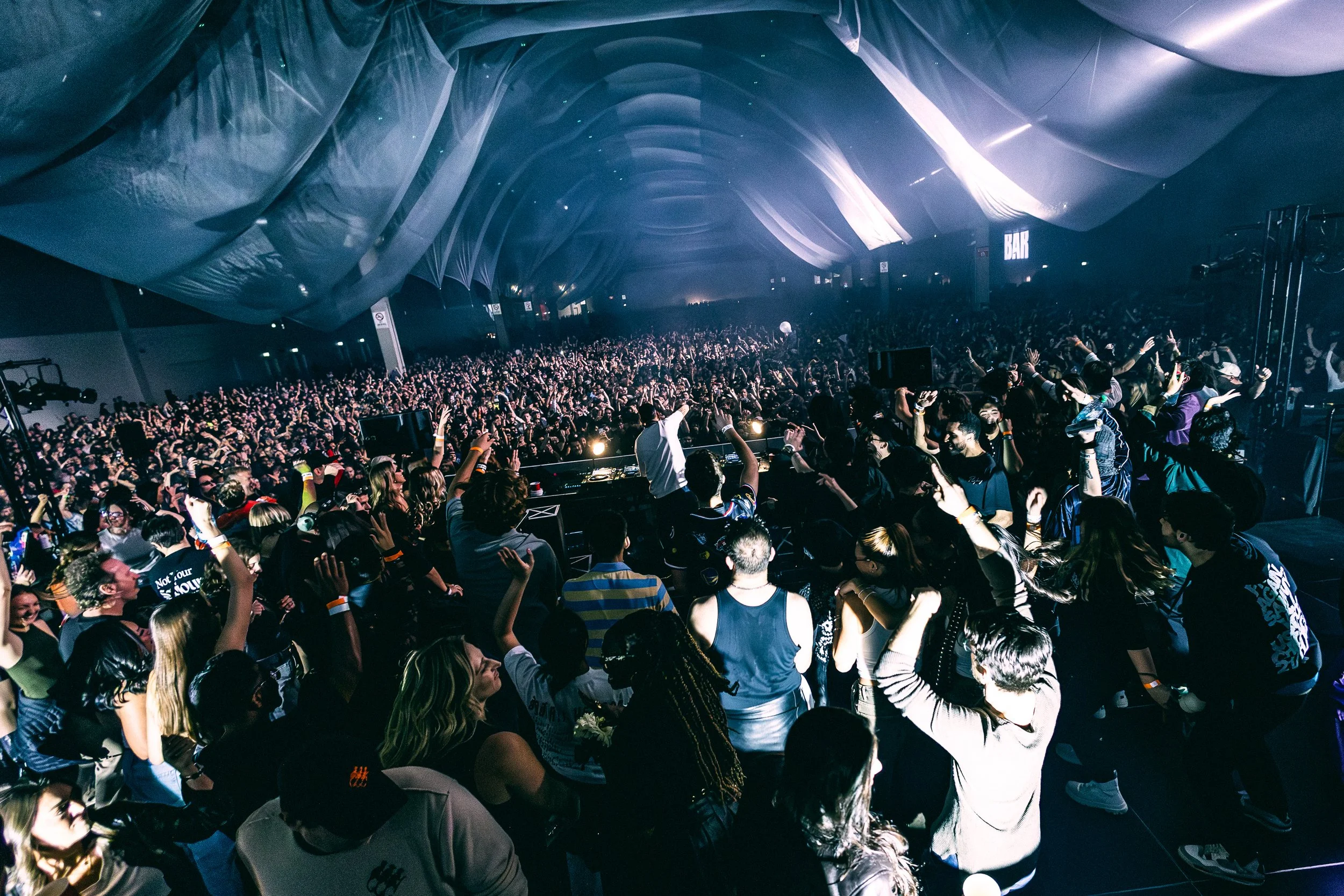 Crowd of people at a concert or music festival in a large indoor venue with a fabric ceiling, cheering and dancing.