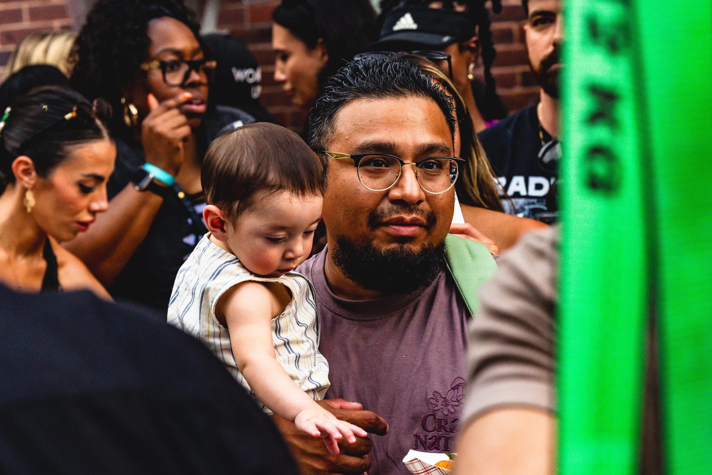 A man with glasses and a beard holds a young child with a bowl-cut hairstyle on his shoulder. The child is looking down at something in his hand. Several women and men are visible in the background, some wearing black clothing and glasses, with a bri