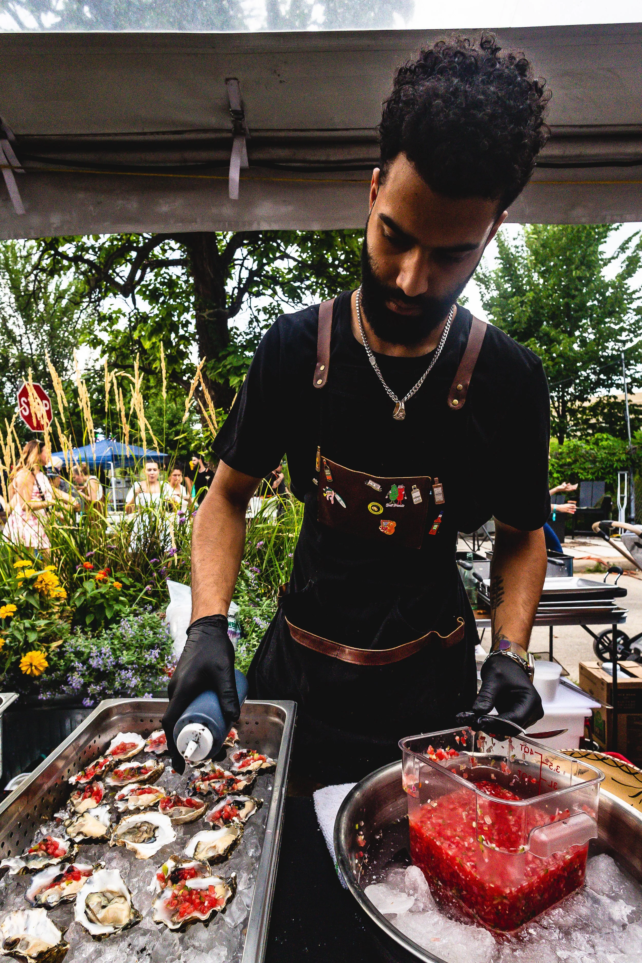 A man wearing black gloves and a black apron is preparing oysters with a topping at an outdoor event.