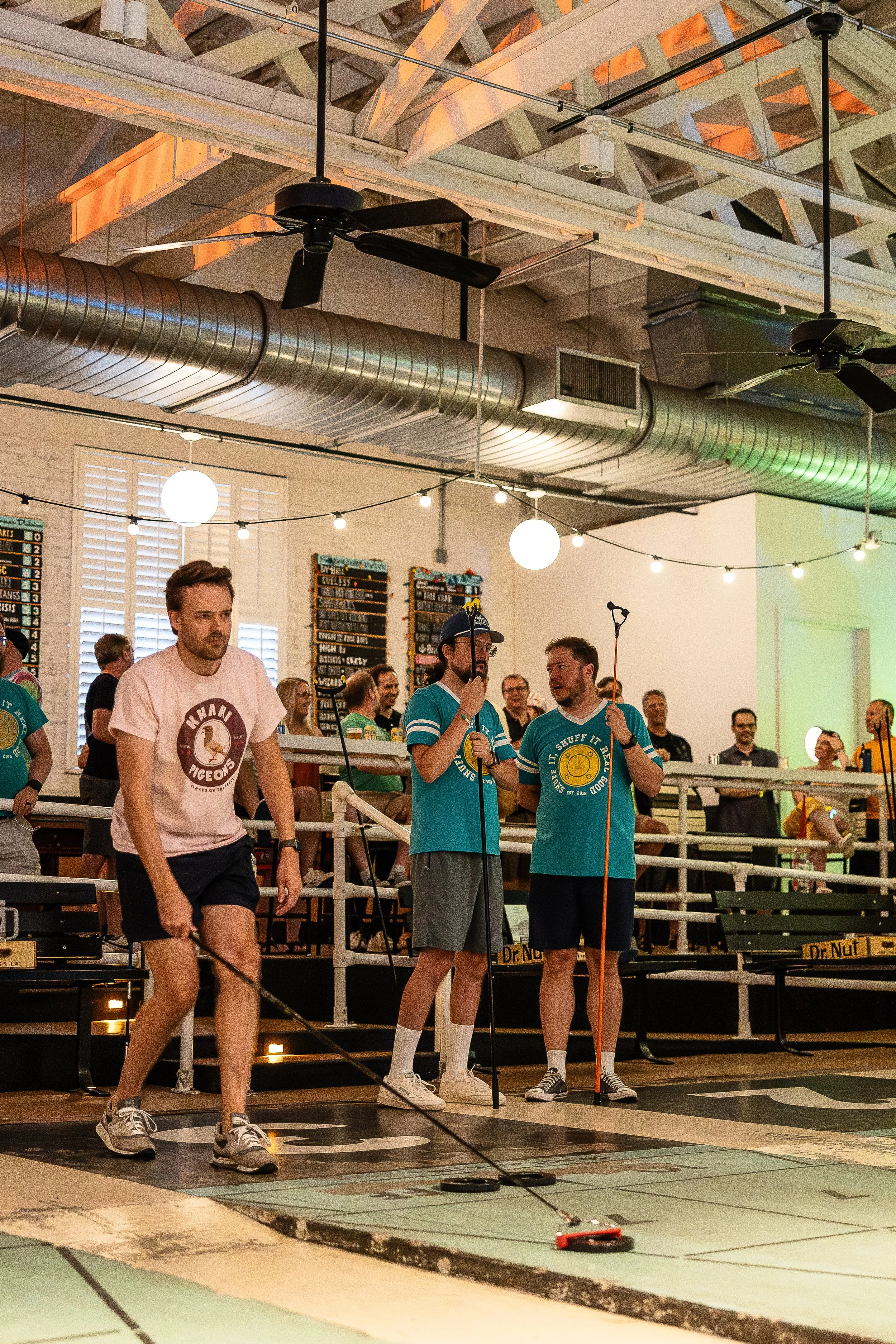 A group of people playing shuffleboard indoors. One person is sweeping the court while others are holding shuffleboard cues. There are spectators in the background watching the game.