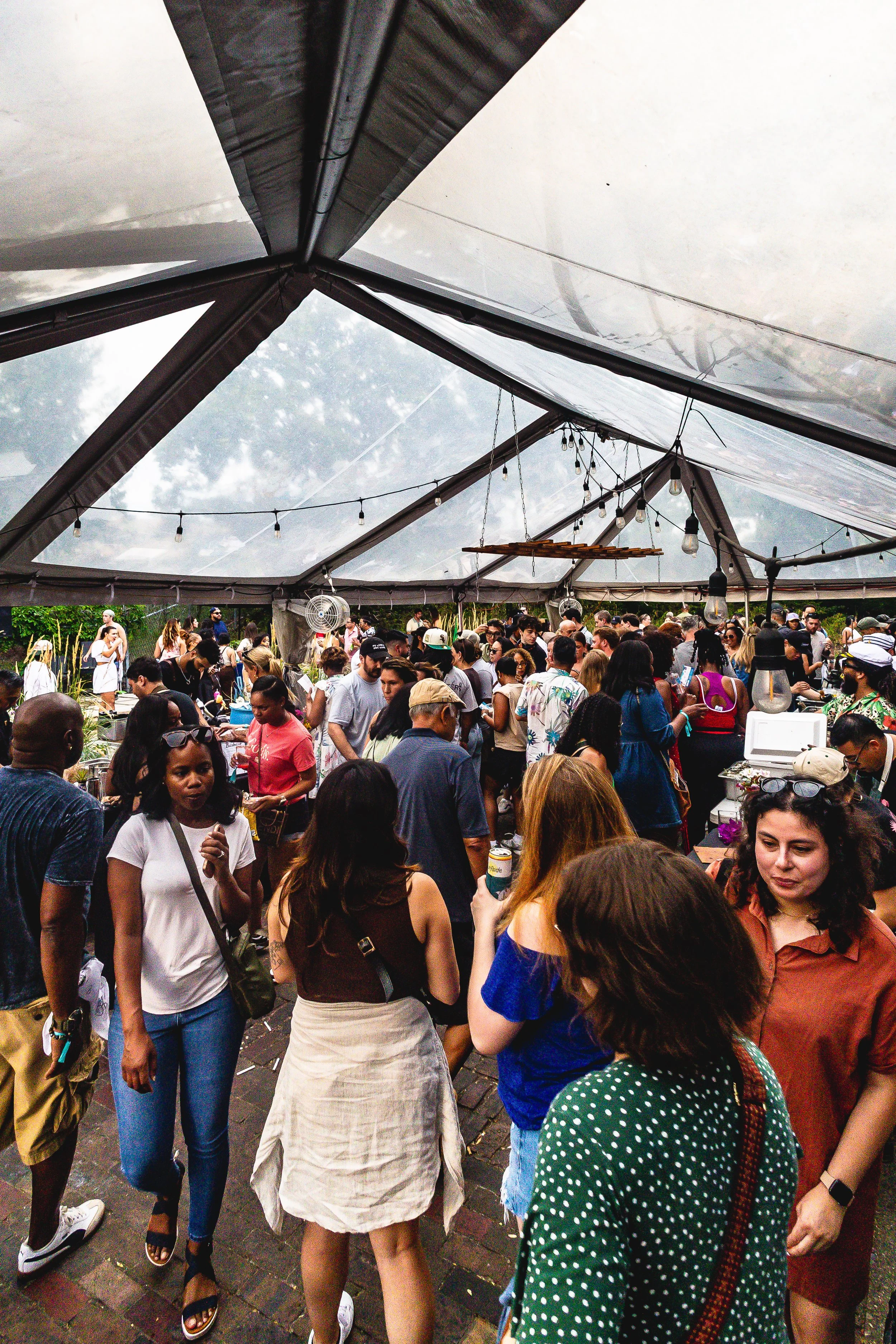 Crowd of people at an outdoor event under a large tent with string lights and fans.