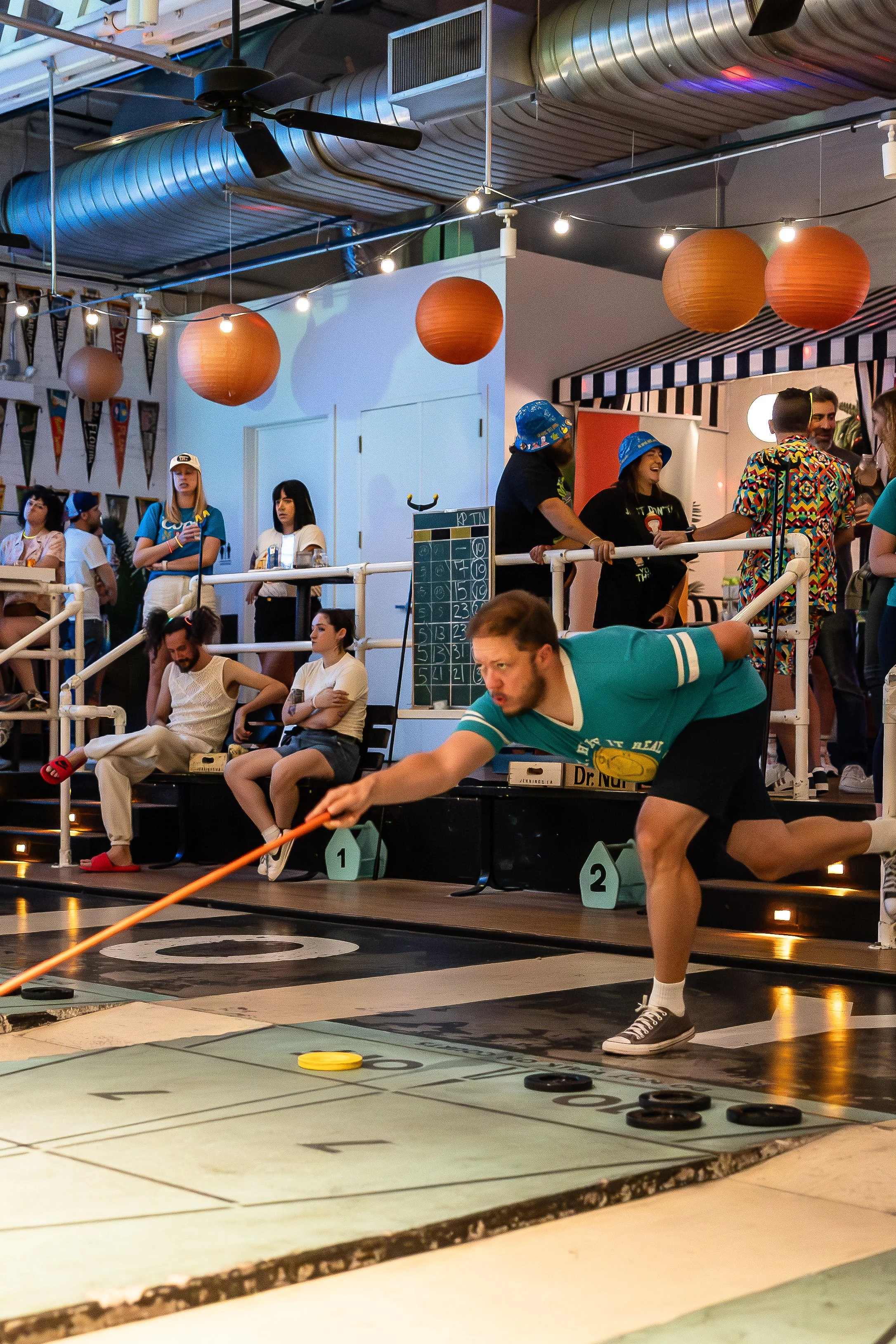 Man playing shuffleboard at an indoor recreational space with colorful lighting and hanging paper lanterns, surrounded by spectators and other players.