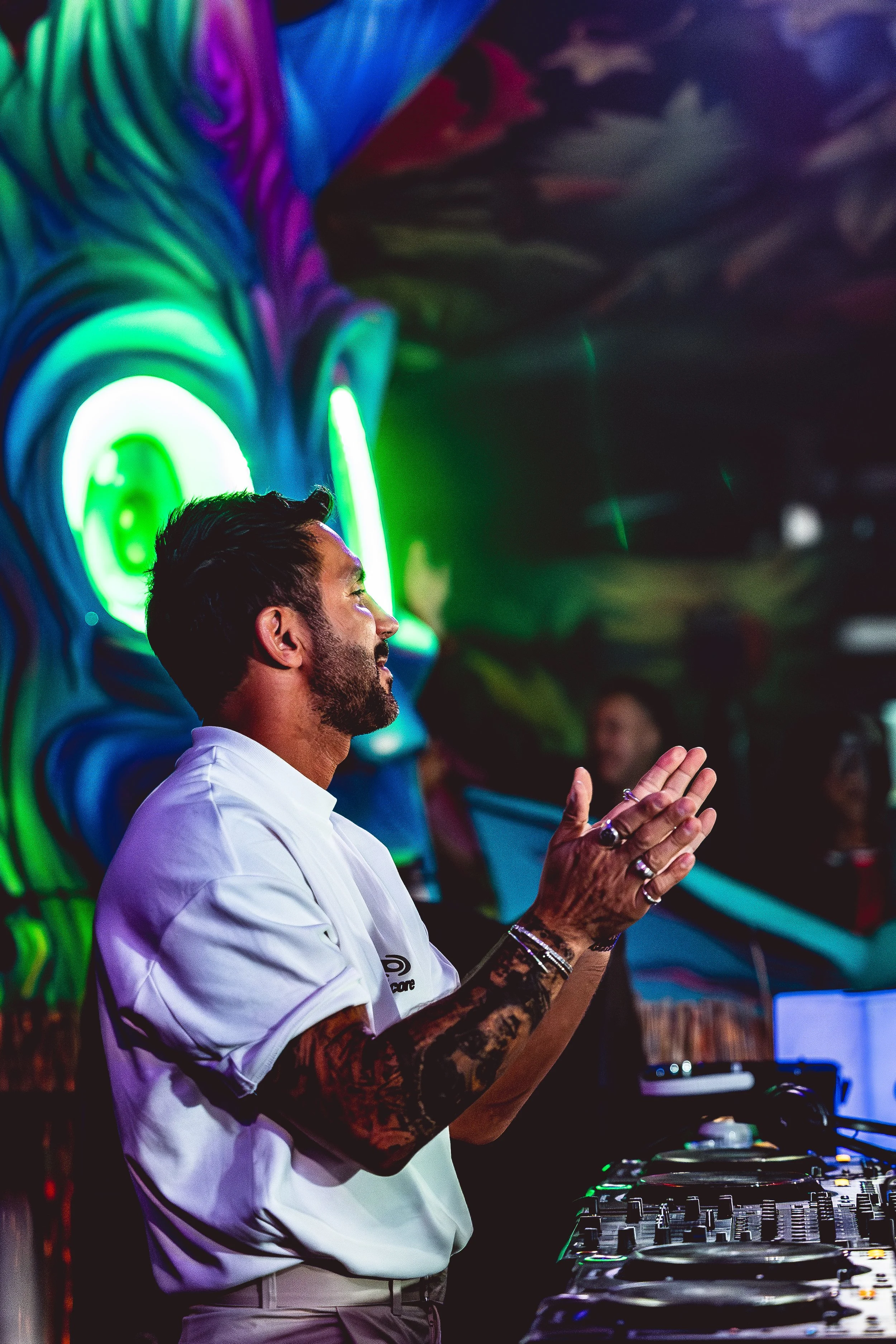 A man with a beard, tattoos, and rings on his fingers is DJing at a colorful event, with neon lights and a mural in the background.