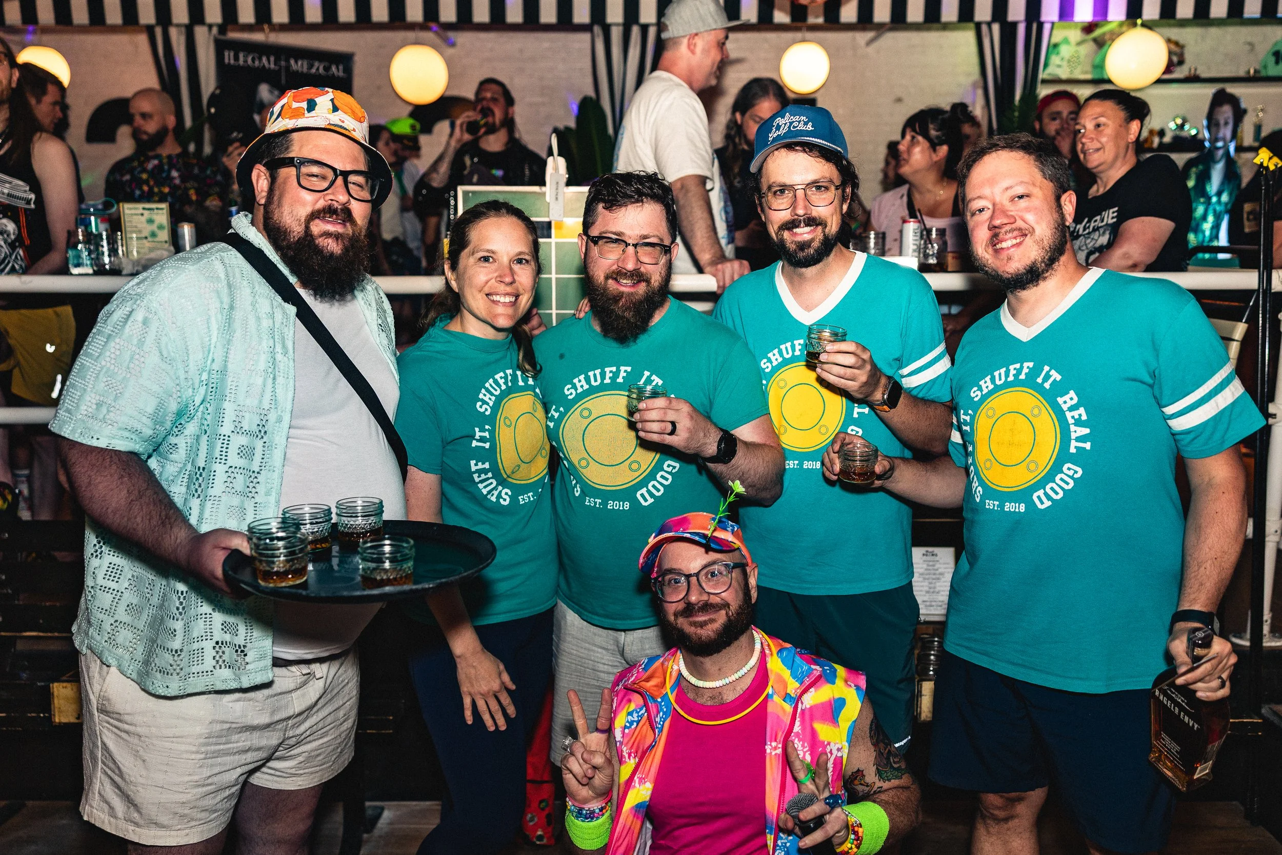 Group of people at a lively social event, some wearing matching teal shirts that read 'It's SHUFF IT REAL GOOD,' with a man in the front wearing colorful, flashy clothing and a hat, holding a peace sign and a microphone, celebrating in a bar-like set