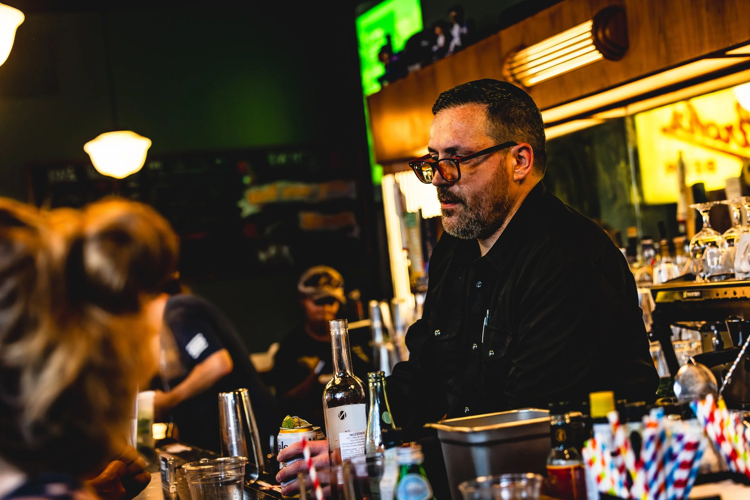 A bartender with glasses and a beard working at a bar counter, serving drinks to customers. The bar has various bottles and glassware, with warm lighting and a vibrant atmosphere.