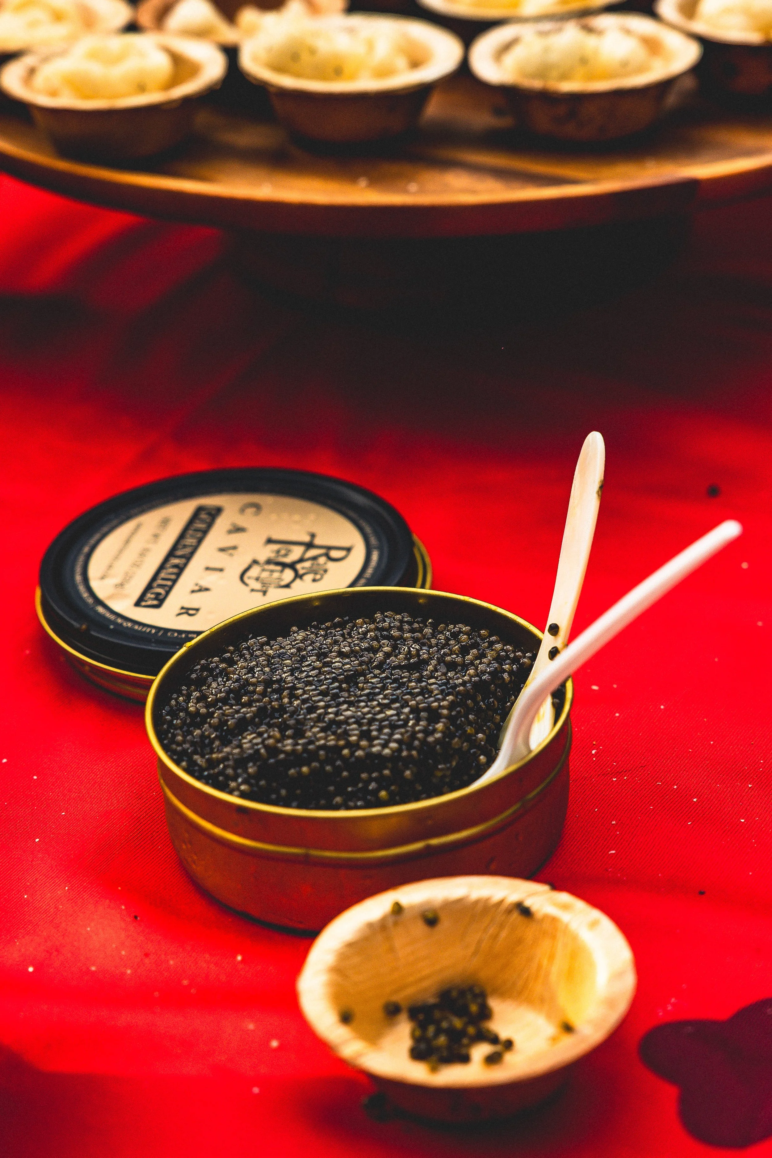 A container of black caviar with two small spoons and a small bowl of caviar in front, with a wooden tray of small dishes in the background on a red tablecloth.