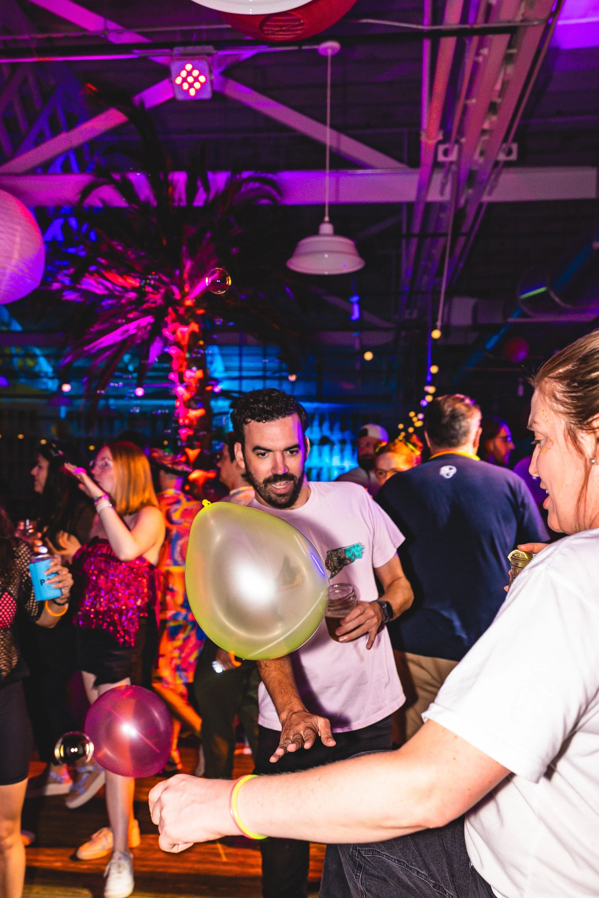 People dancing at a party with neon lights, balloons, and a decorative palm tree in the background.