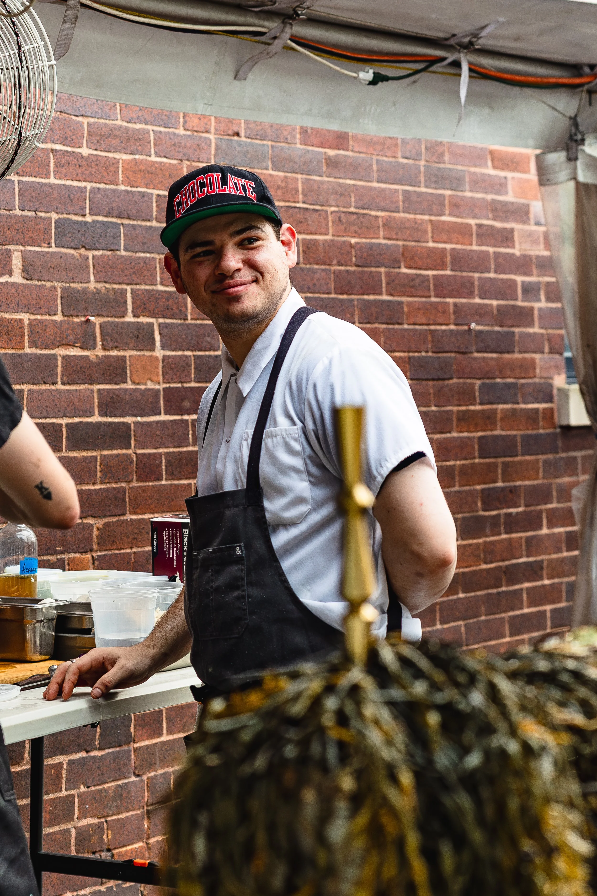 A young man in a white shirt, black apron, and a black cap with red and white text, smiling and standing at a food stall with a brick wall in the background.