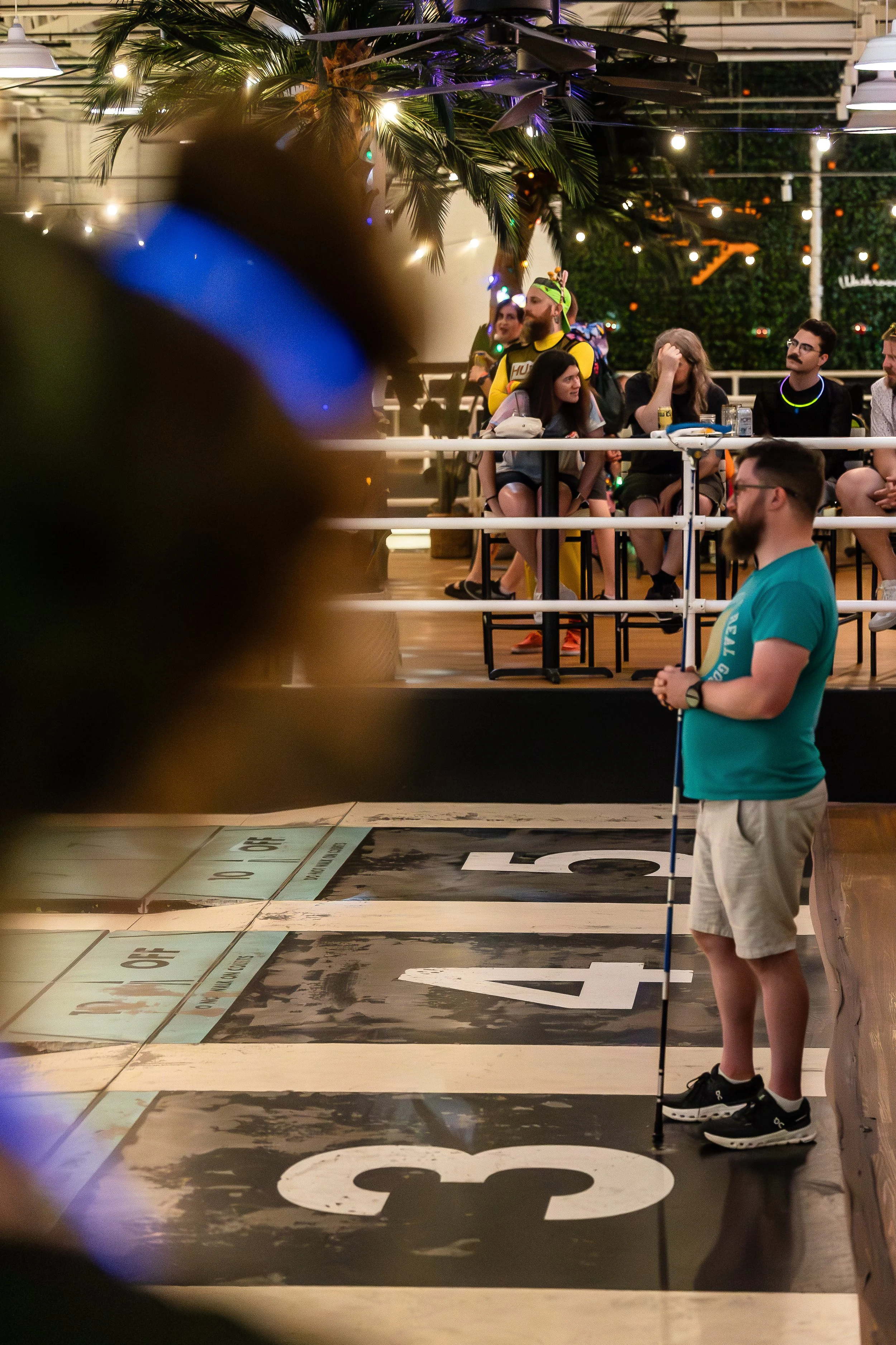 A man with a beard, wearing glasses, a turquoise t-shirt, beige shorts, and sneakers, standing next to a golf club on a miniature golf course at night. In the background, a group of people sitting at a bar with illuminated palm trees and string light