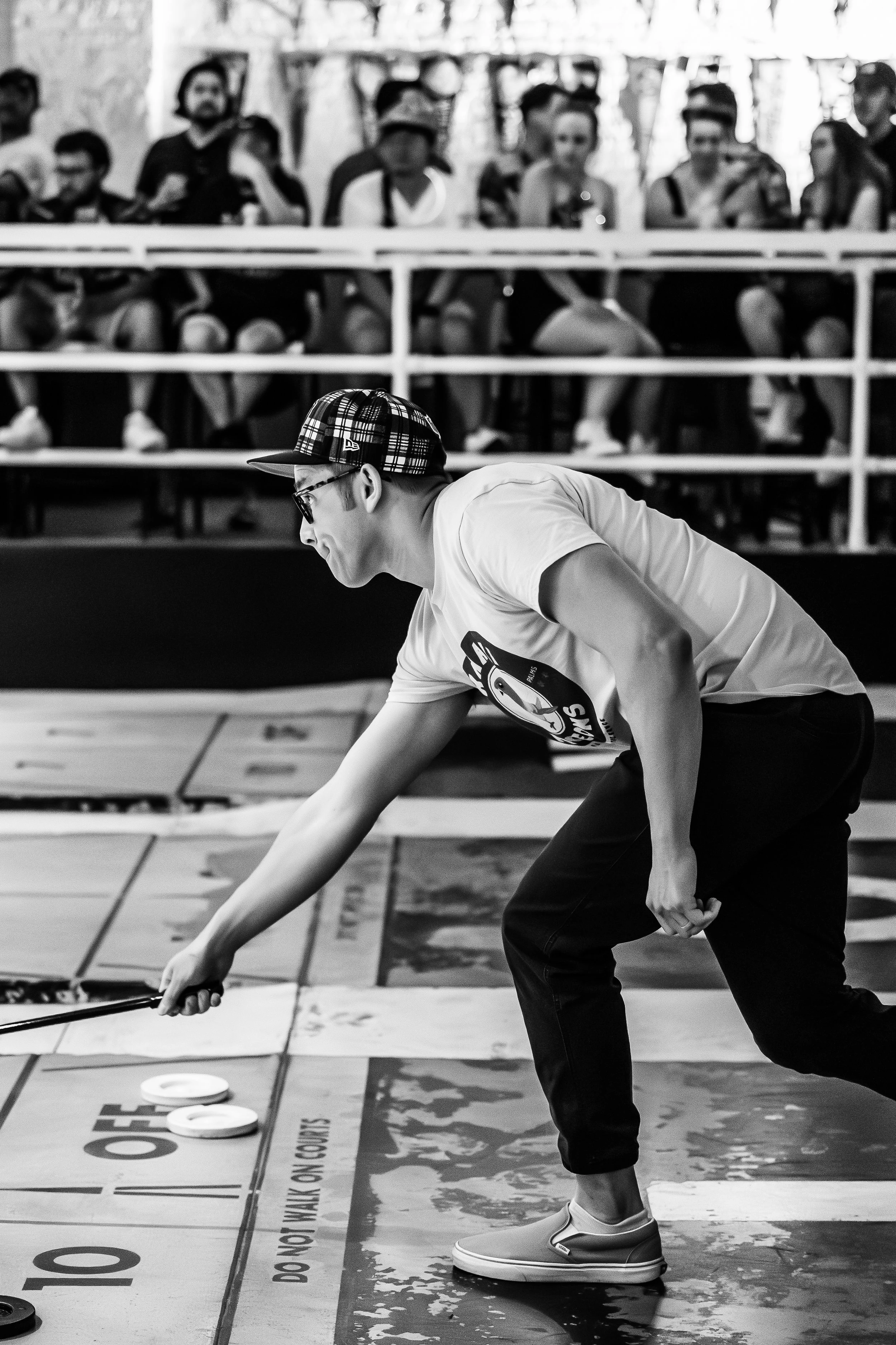 A young man in a baseball cap, glasses, and casual clothing playing shuffleboard in front of an audience at an indoor venue.
