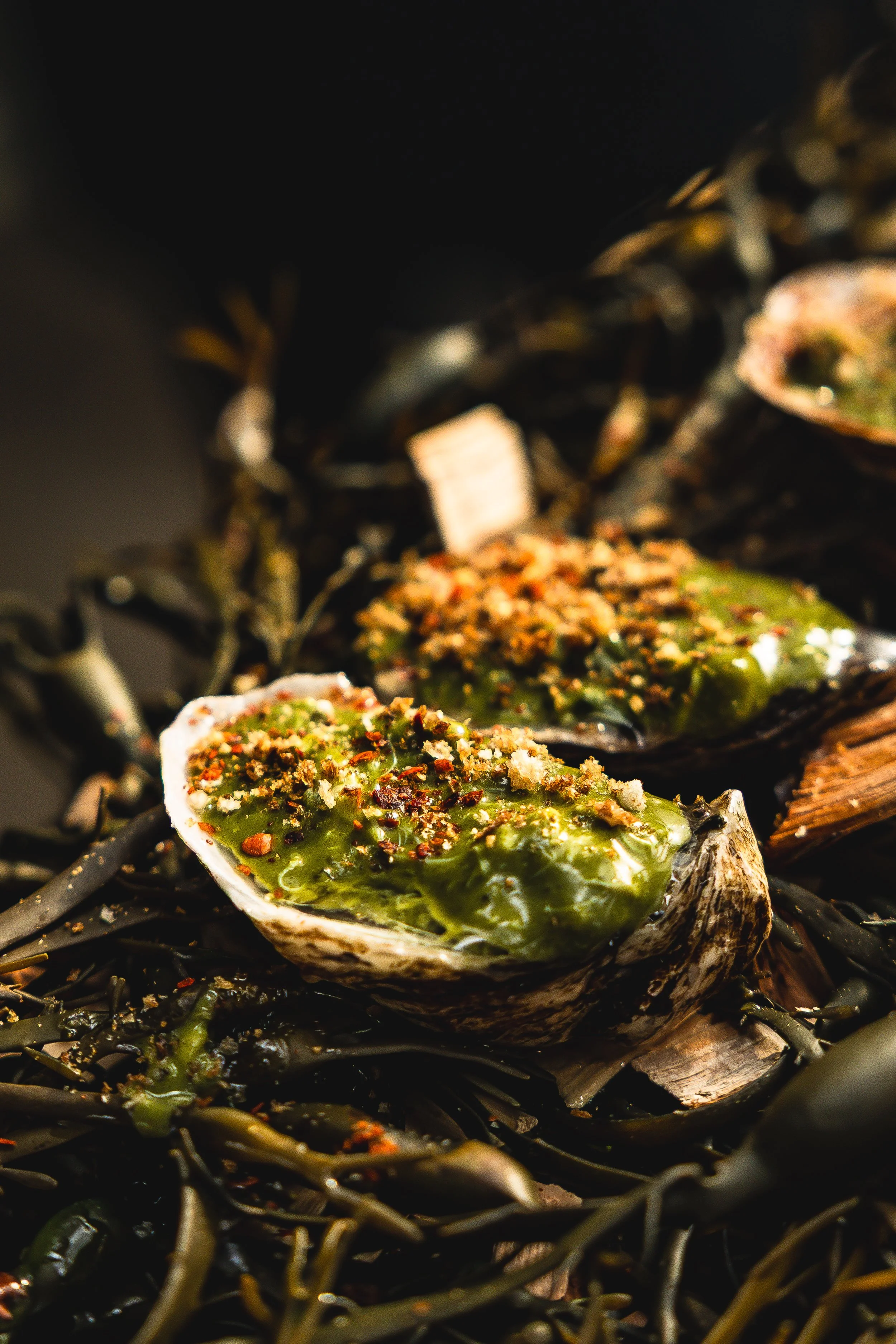 Close-up of cooked oysters topped with green sauce and red pepper flakes on a bed of dark seaweed.