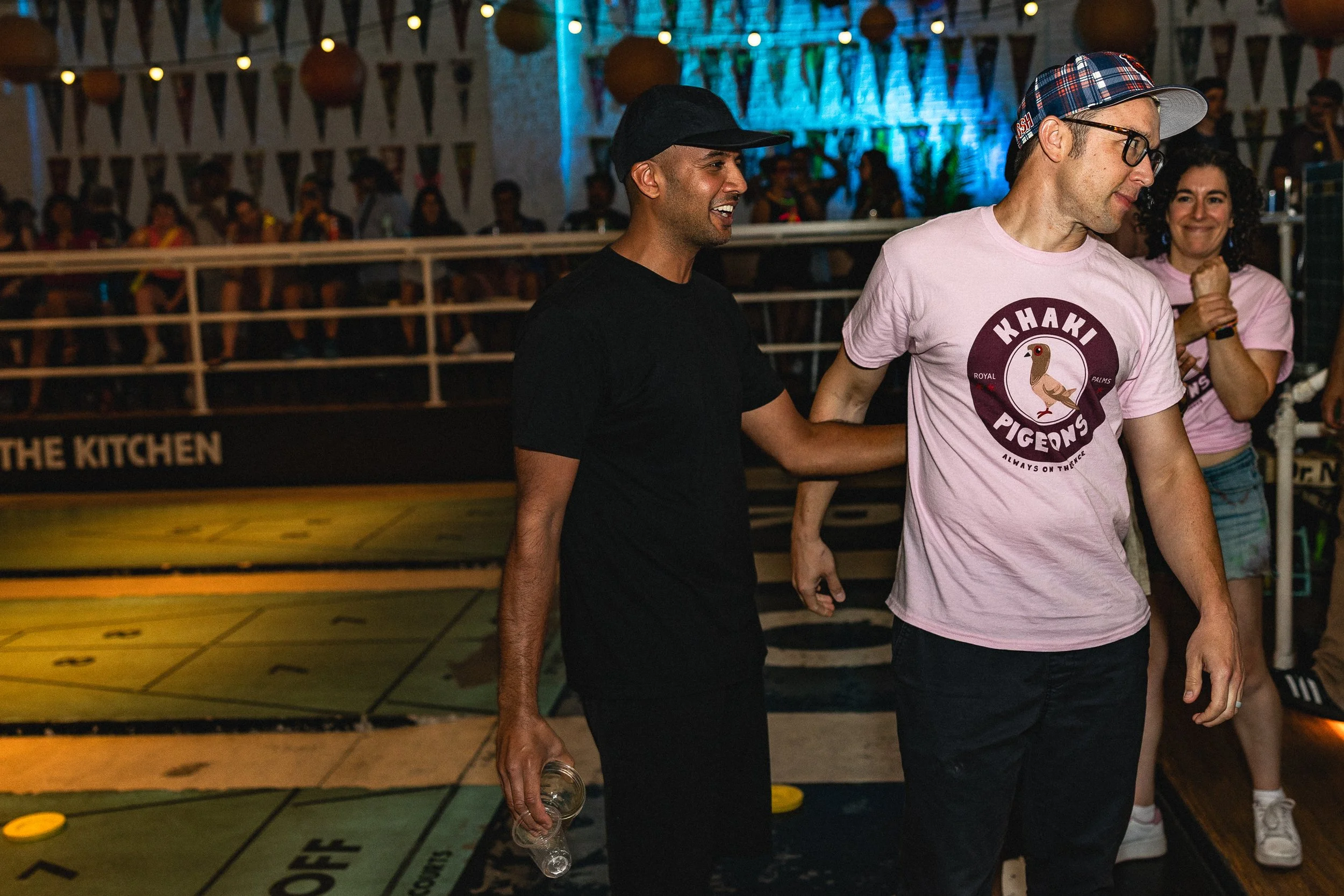 People playing arcade shuffleboard in a lively indoor setting with decorative flags and string lights.