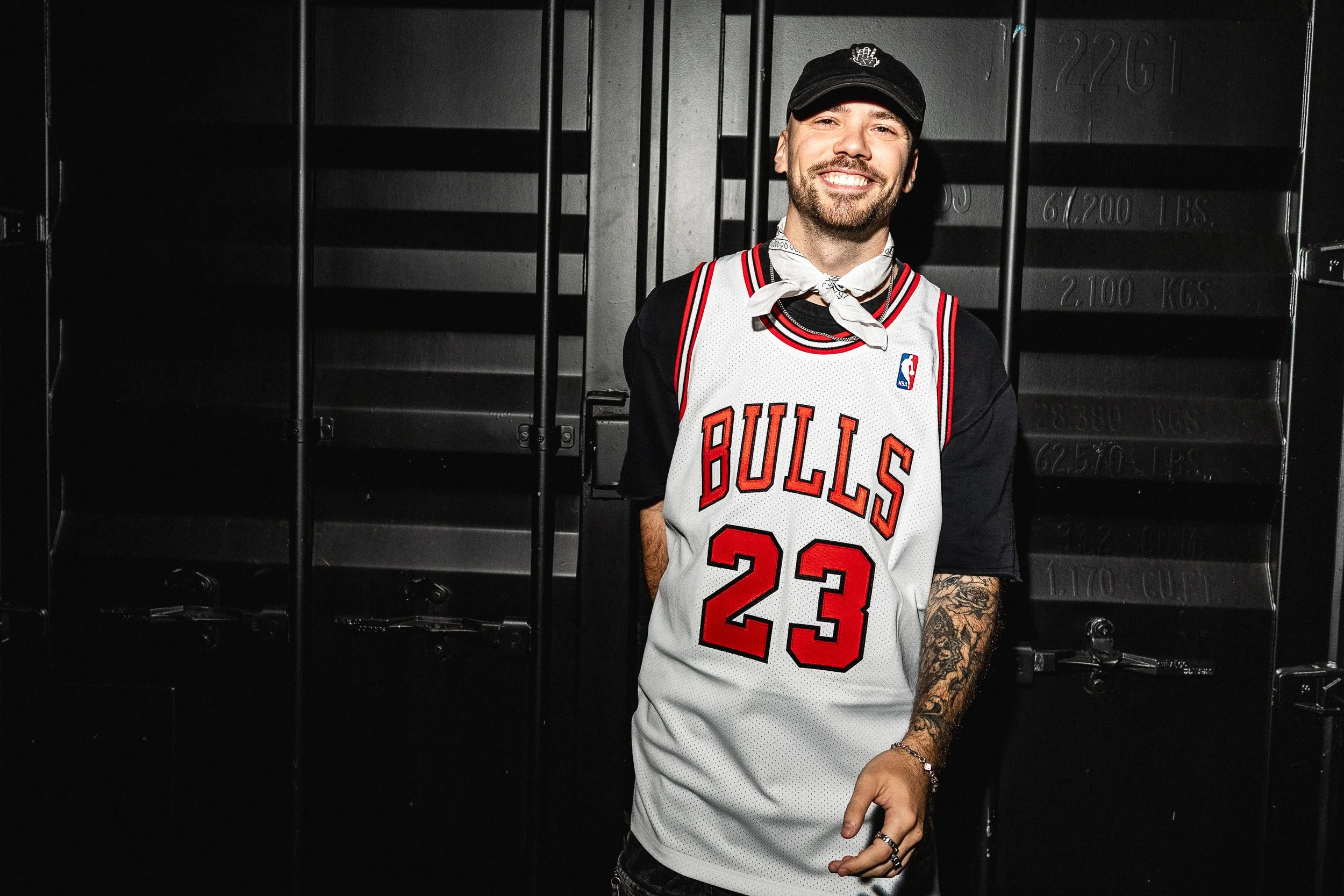 A smiling man wearing a Chicago Bulls basketball jersey with the number 23, black T-shirt, black cap, and a white bandana around his neck, standing in front of a black locker.