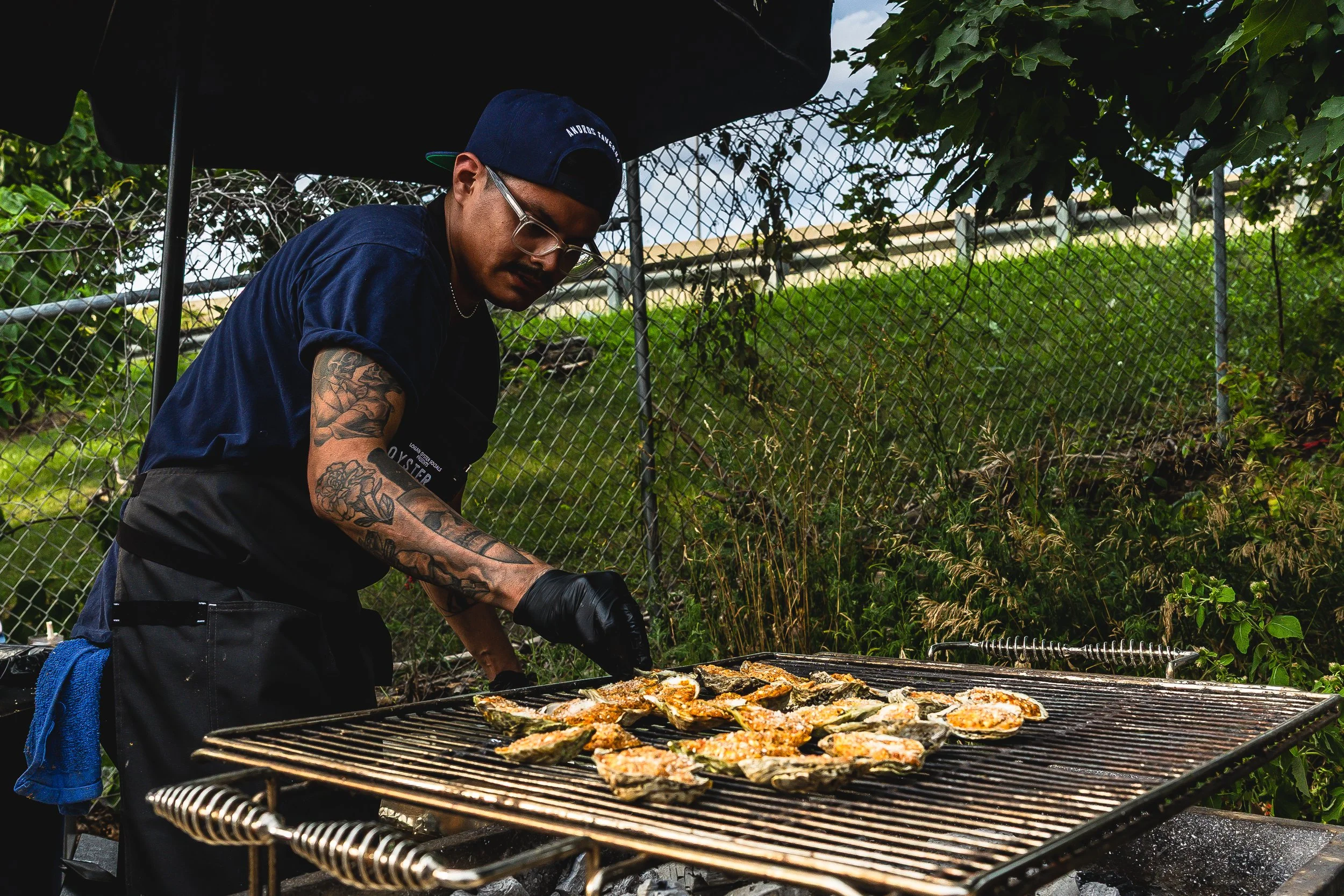 A man with tattoos on his arm wearing a blue cap, glasses, black gloves, and a navy T-shirt is grilling oysters on a barbecue grill outdoors.