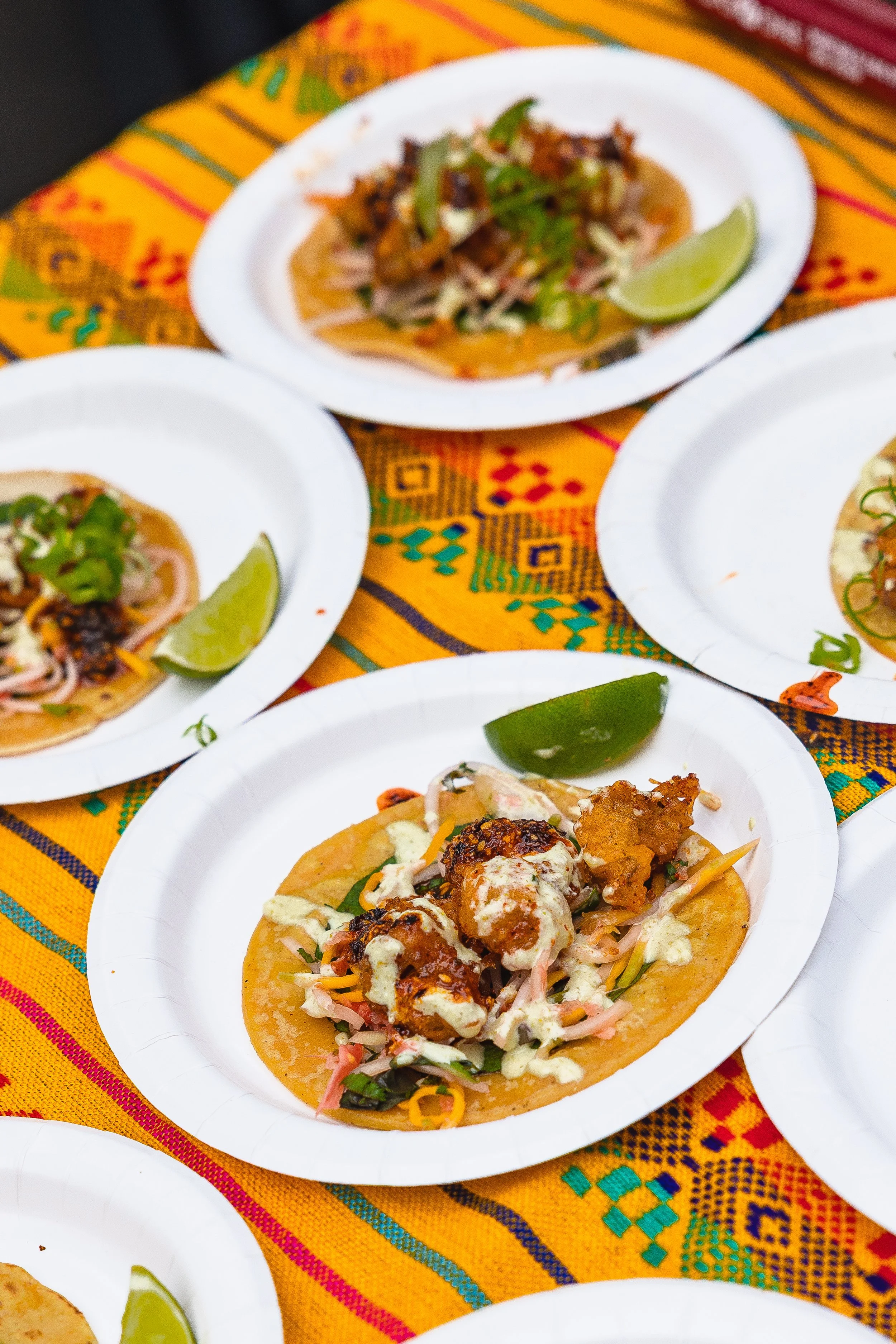 Several plates of tacos with various fillings and garnishes, served with lime wedges, on a colorful, patterned tablecloth.