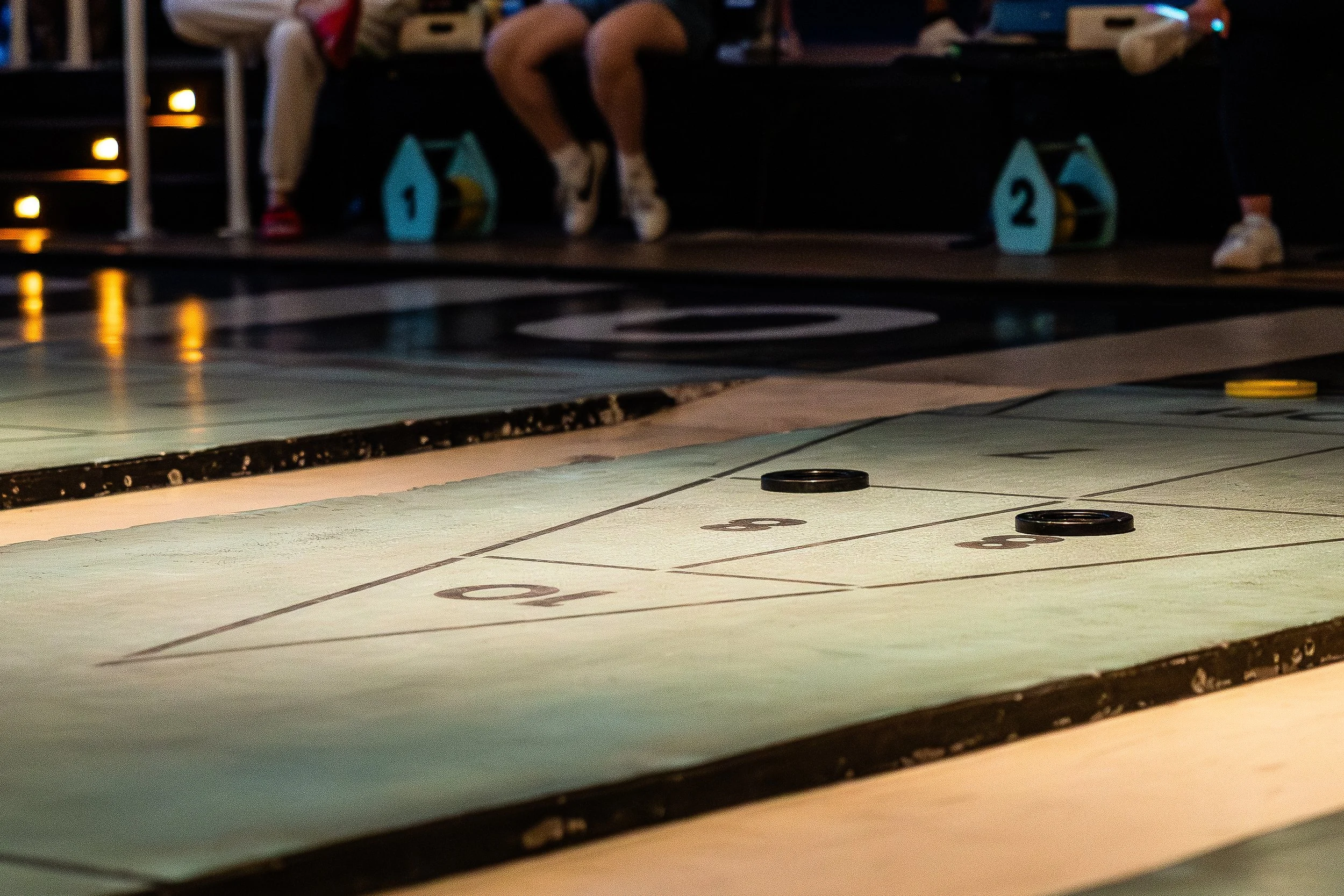 The image shows a shuffleboard court with pucks on the playing surface and players sitting on the bench in the background.