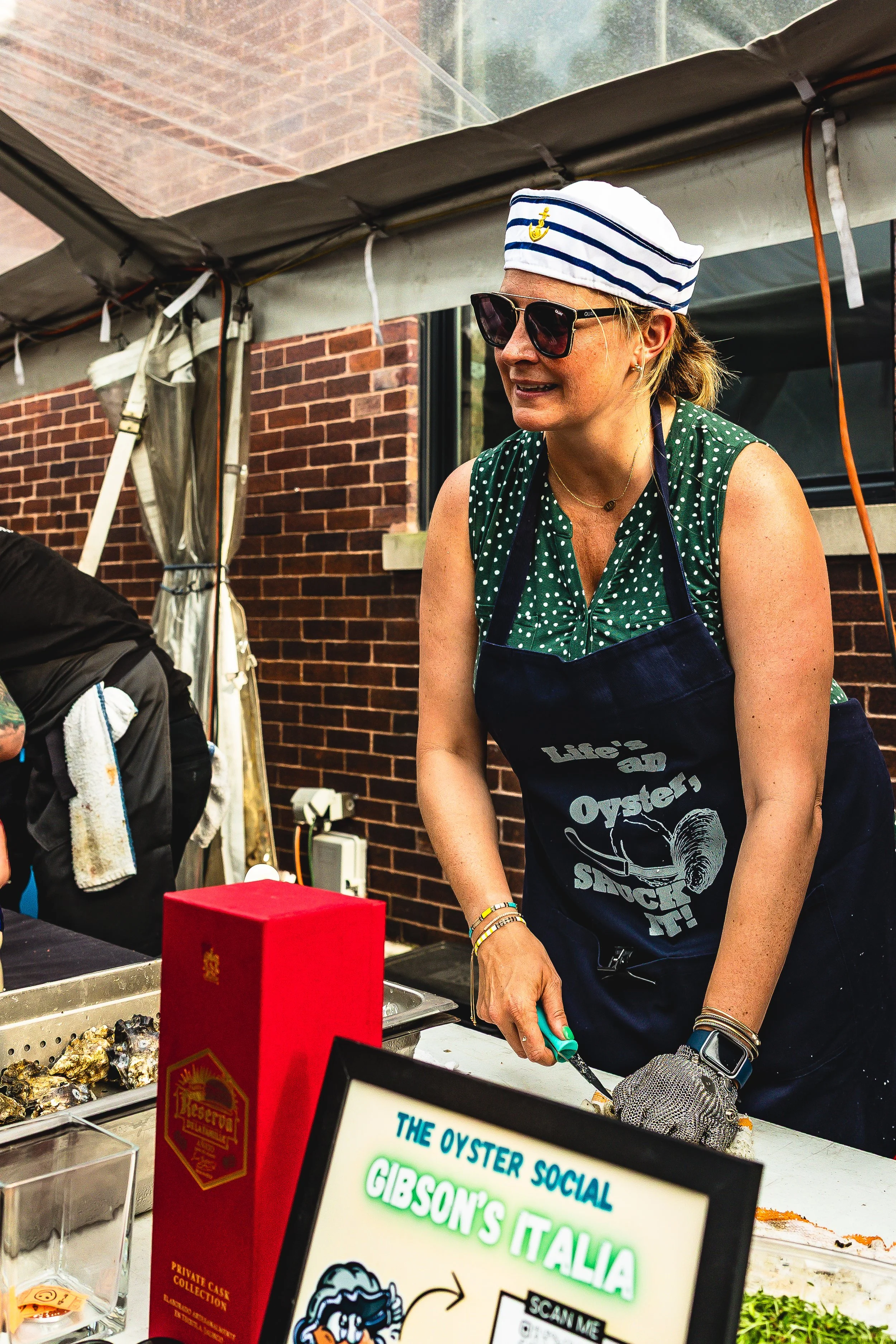 A woman wearing sunglasses, a striped sailor's hat, and a navy apron, preparing oysters at an outdoor seafood stand with a sign reading 'Gibson's Italia'.