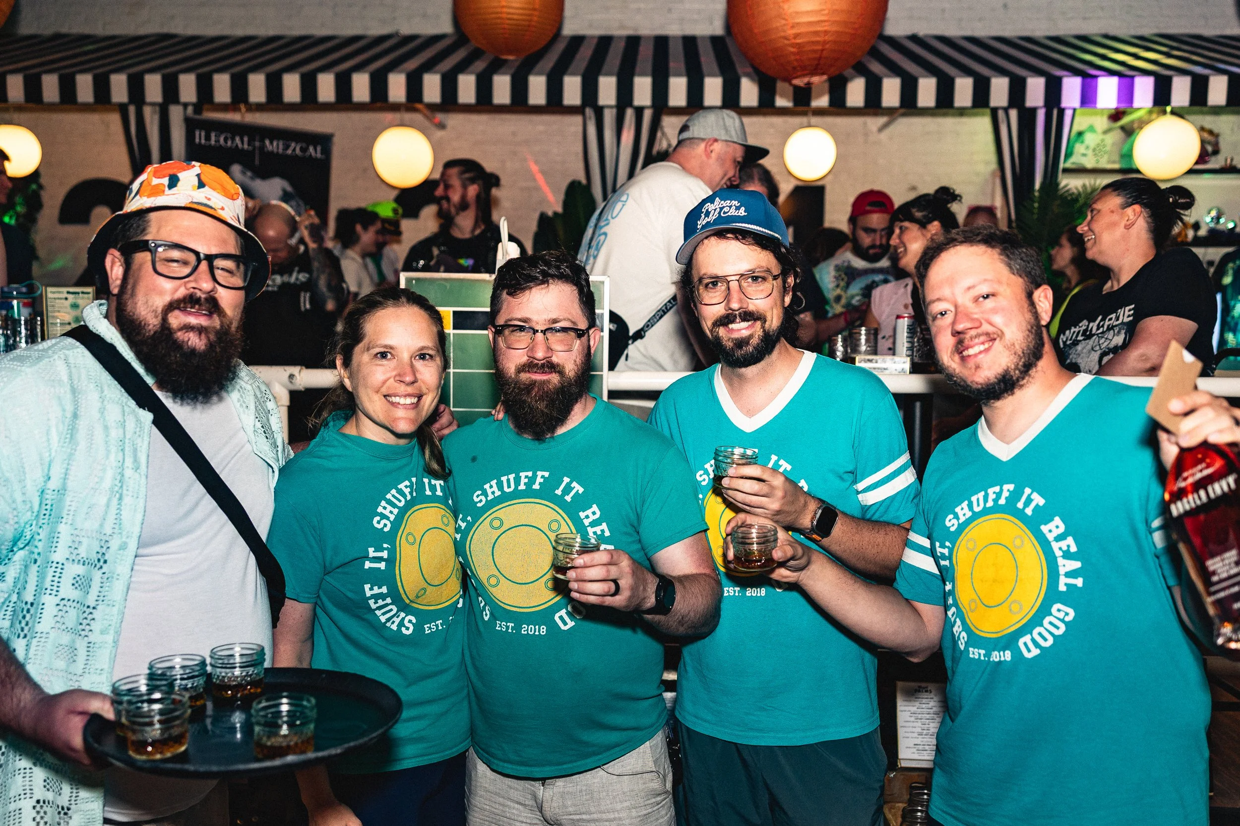Group of five friends in a bar or party setting, smiling and holding glasses of dark liquor. They are wearing matching teal T-shirts with a yellow beer cap logo and the phrase 'IF SHUFF IT REAL,' Est. 2018. The background shows a lively crowd, bar co