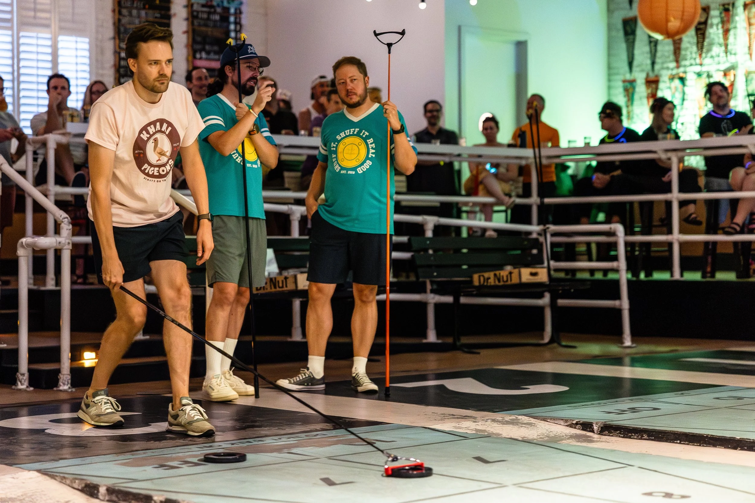People playing shuffleboard on an indoor court with spectators watching in the background.