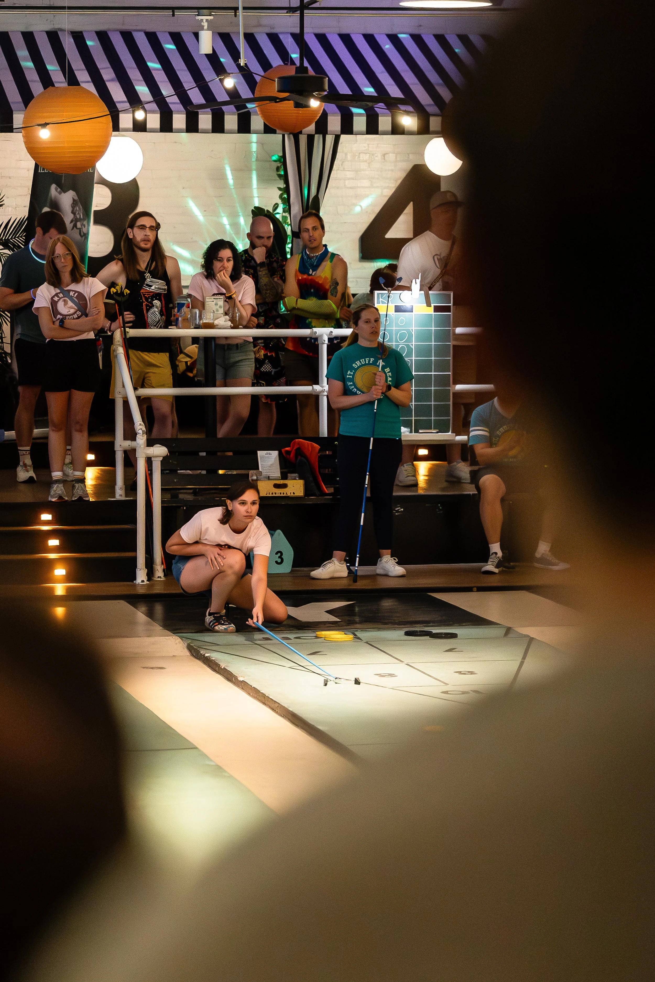 Young woman in white shirt and shorts crouching, playing shuffleboard in a lively indoor venue, with audience and event staff watching behind her.