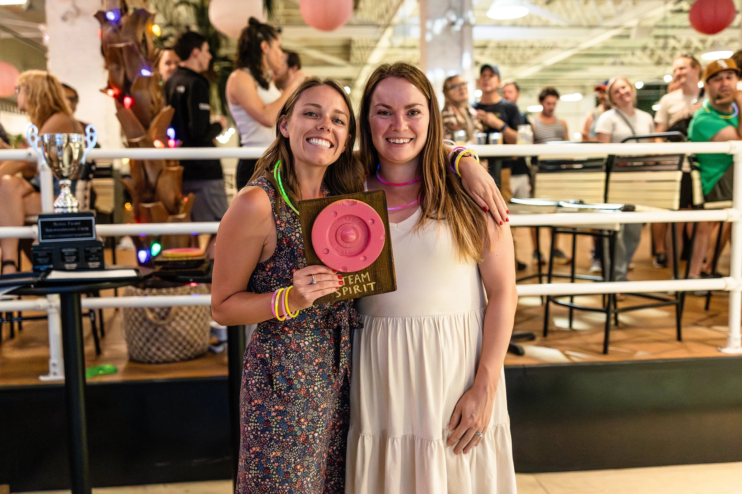 Two women smiling and posing together at a social gathering, one holding a pink speaker award, in a lively indoor setting with other people in the background.
