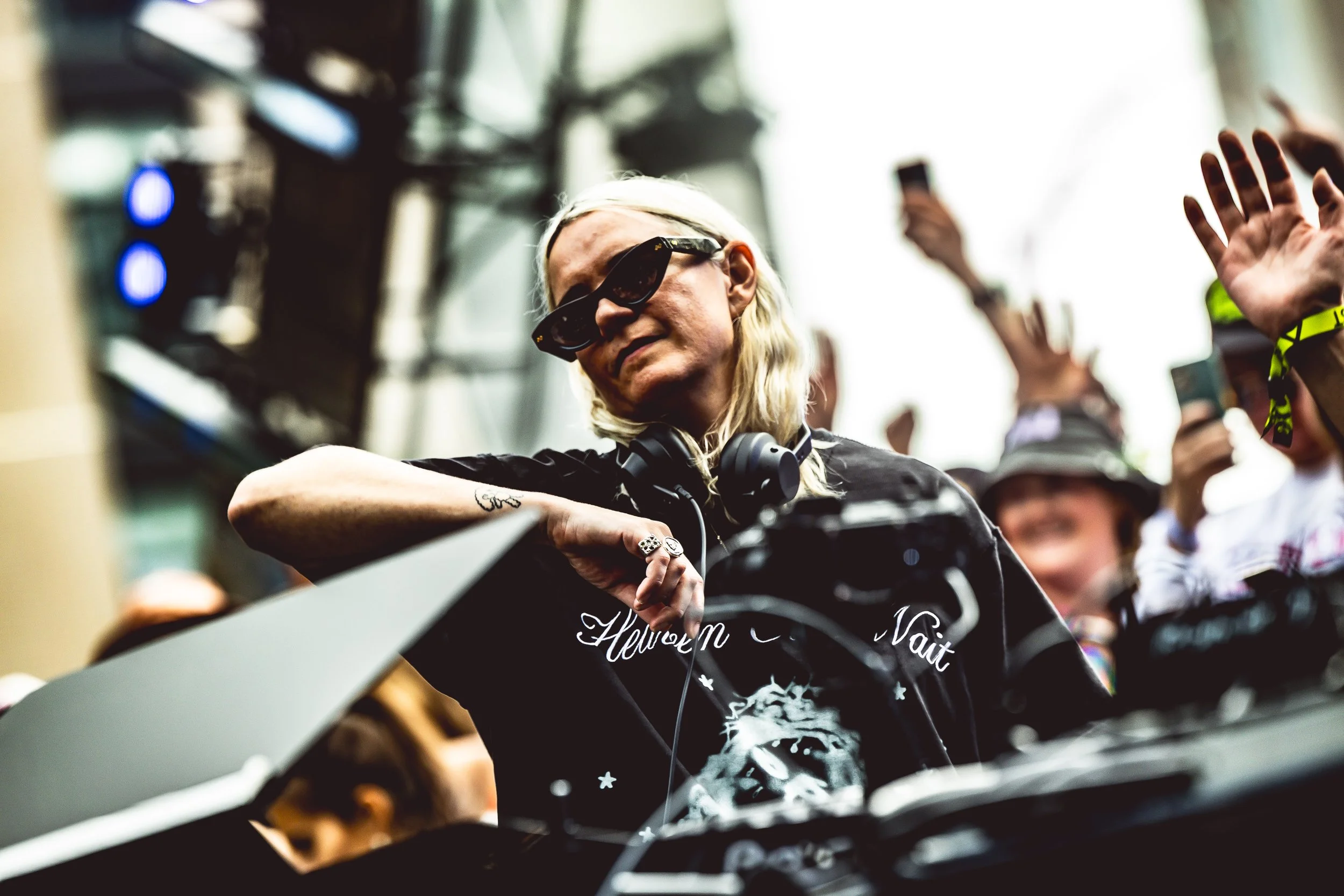 A woman, wearing sunglasses and headphones around her neck, DJing at an outdoor event, with a crowd of people in the background waving and taking photos.