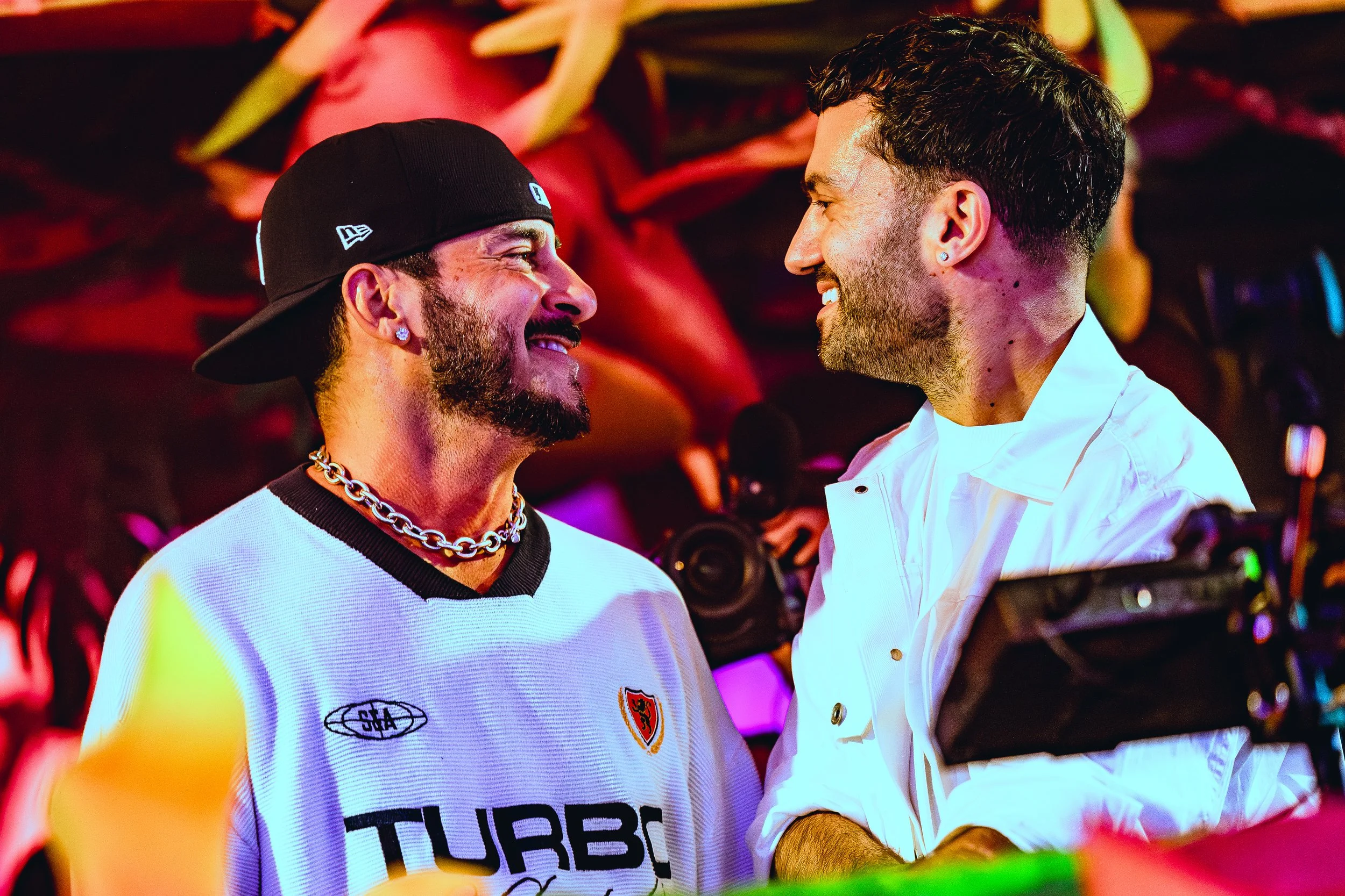 Two men smiling and facing each other in a lively setting with colorful lighting, one wearing a black cap, a white sports jersey with logos, and a chunky chain necklace, the other in a white shirt, both with short dark hair and beards.