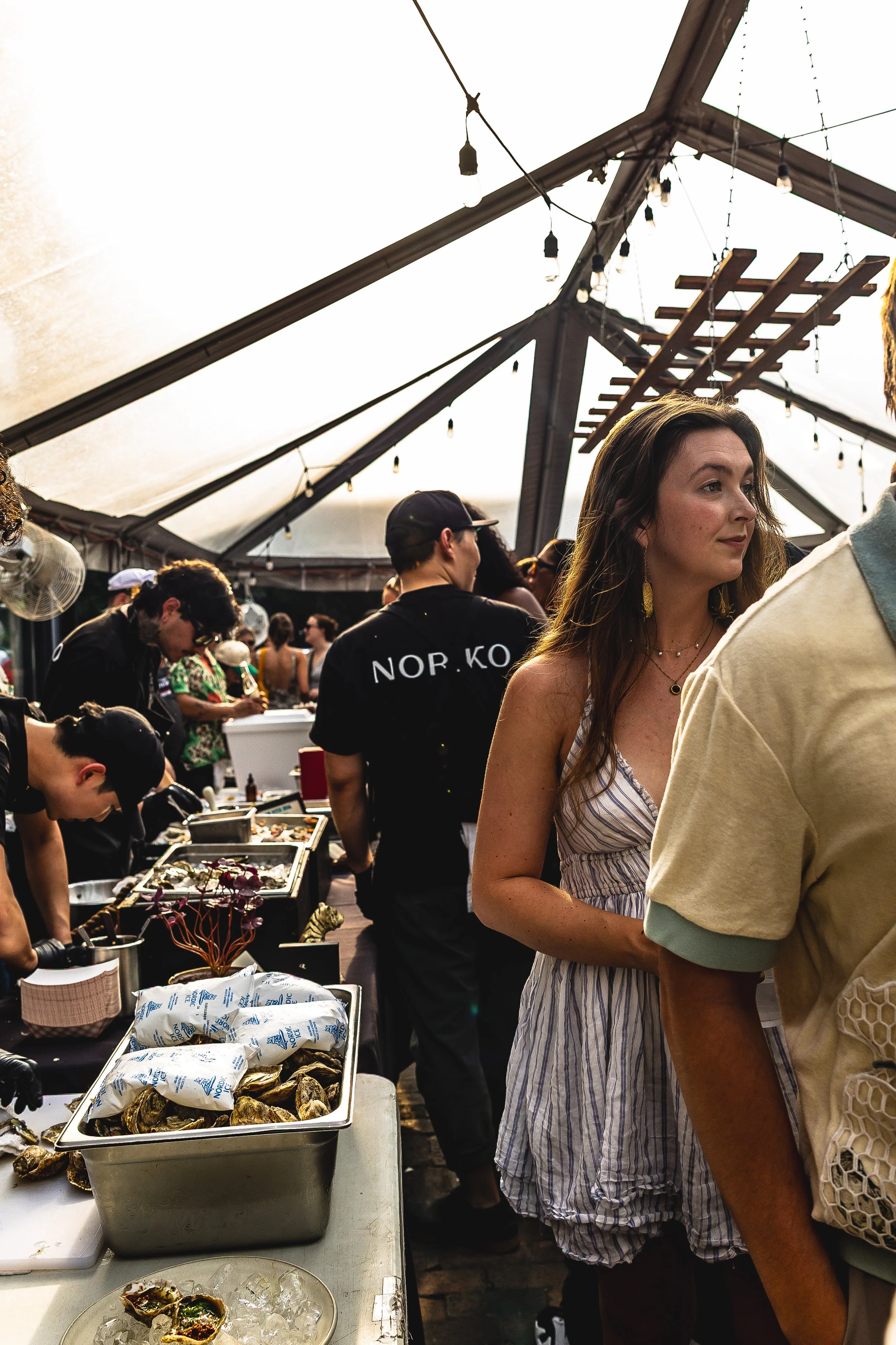 People gathering at a food event under a tent with string lights, with a woman in a striped dress in the foreground and various food trays on a table.