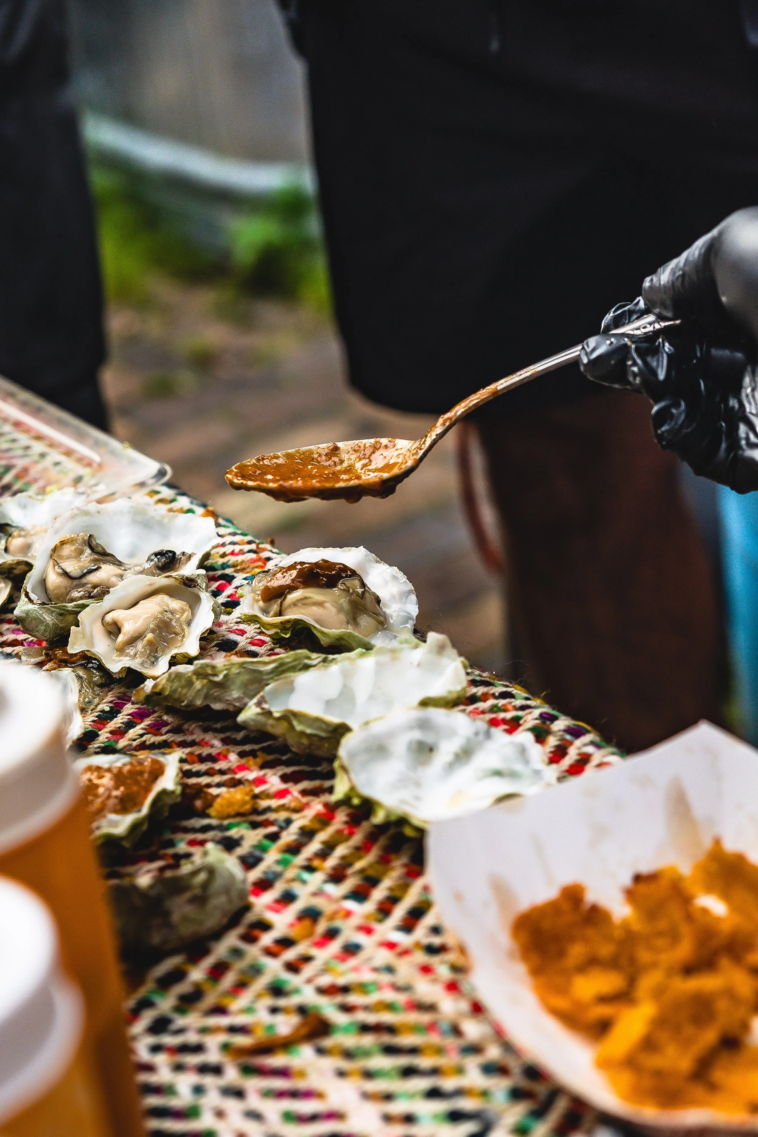 A person wearing black gloves serving sauce over freshly shucked oysters on a colorful woven cloth at an outdoor market.