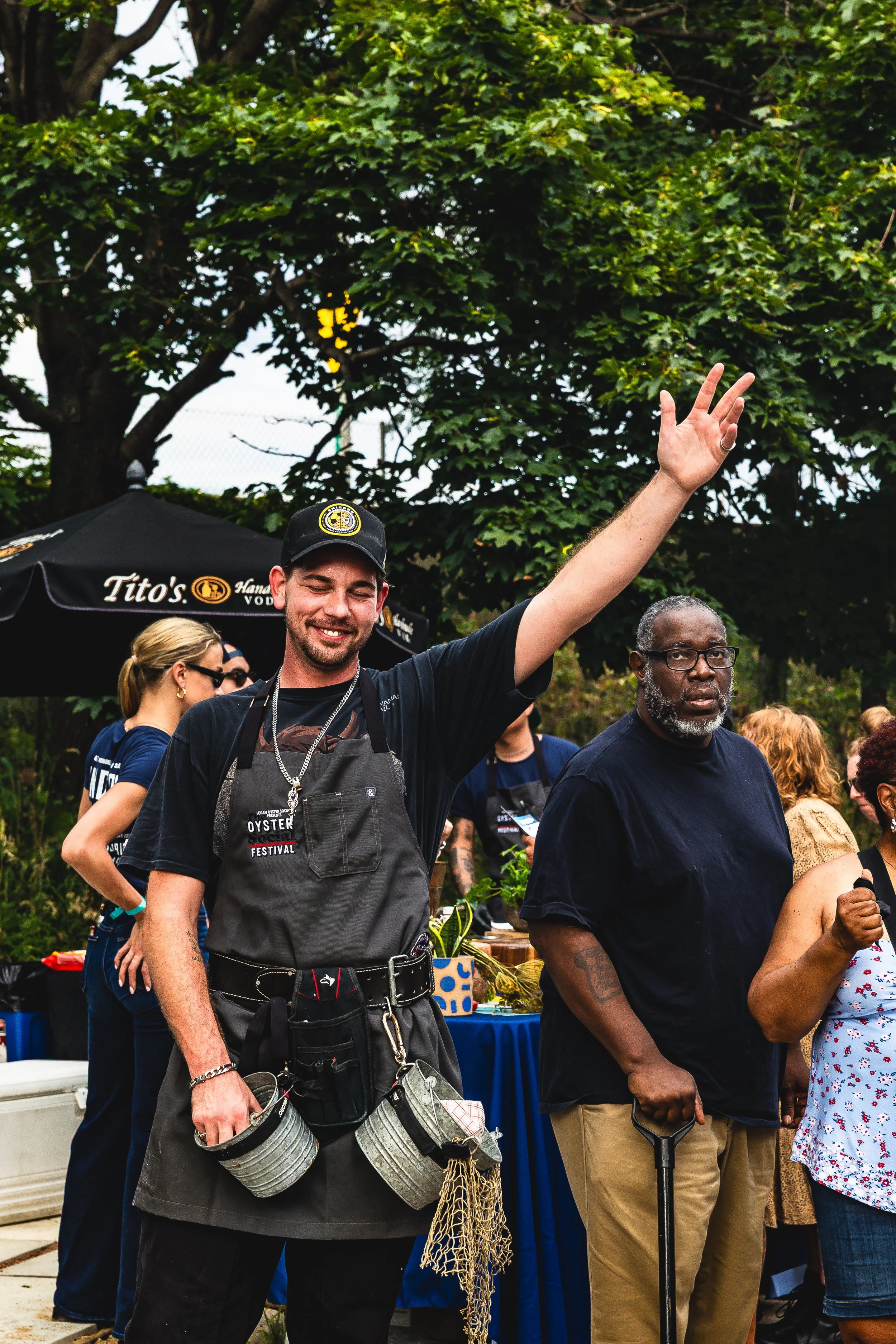A group of people gathered outdoors at a food festival, with a smiling man in the foreground waving and wearing a black cap and apron, next to an older man with glasses holding a cane, and others in the background near a table with food and drinks.