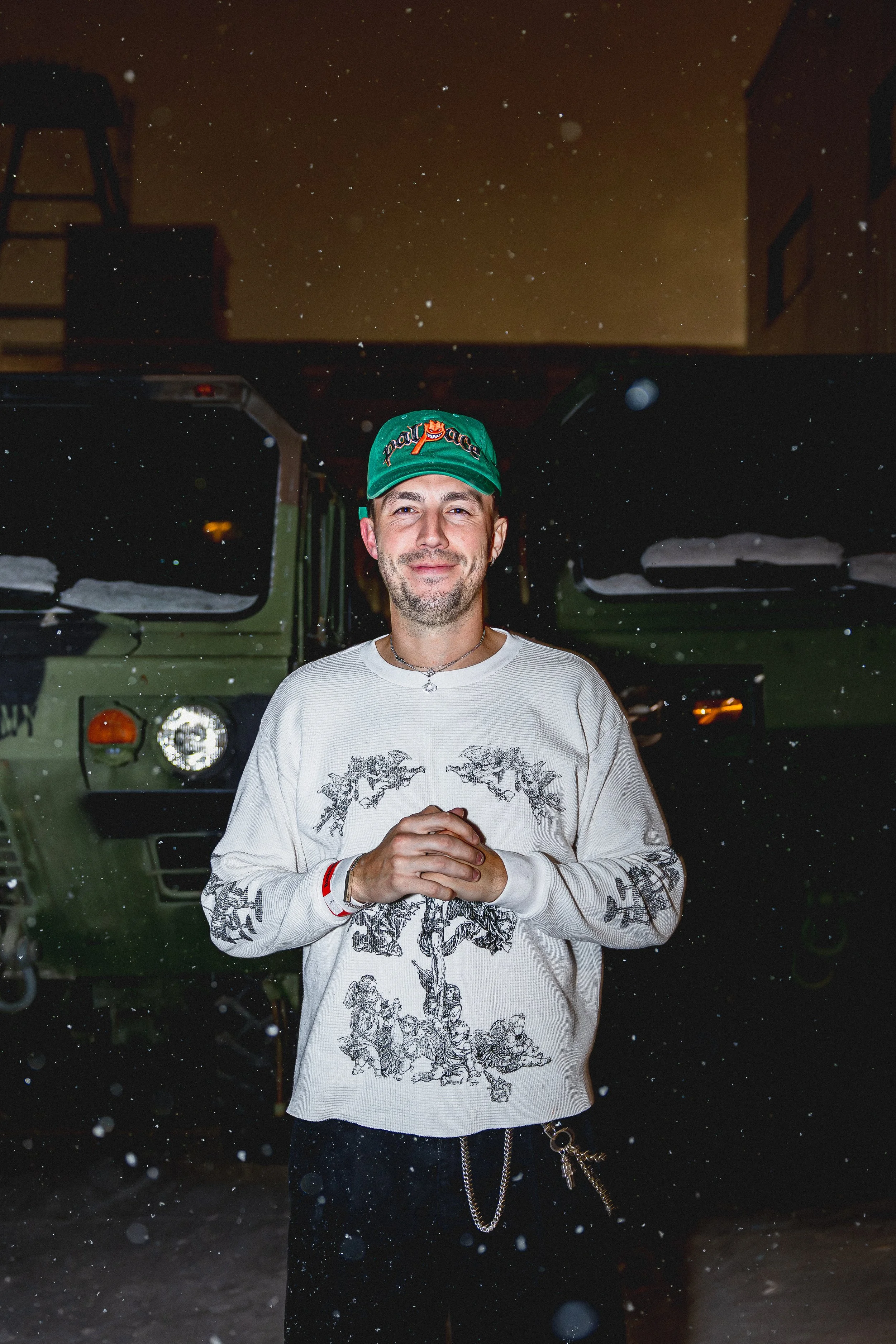 Cloonee with a slight smile, wearing a green cap with orange writing, a white sweatshirt with black illustrations, and chains, standing in front of two green military vehicles at night in snowy weather.