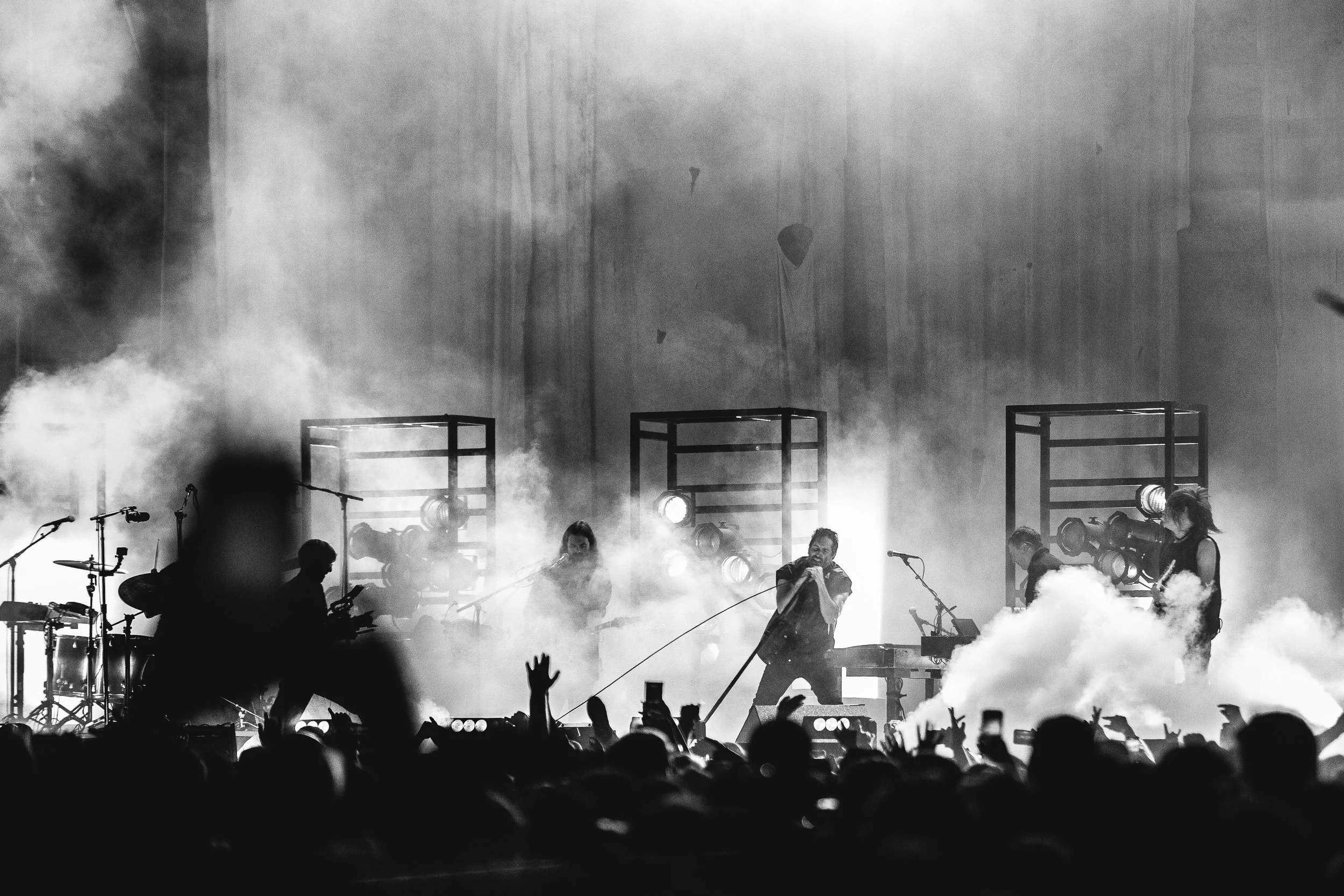 Black and white photo of a live concert with a band performing on stage surrounded by fog, with audience members visible in the foreground.