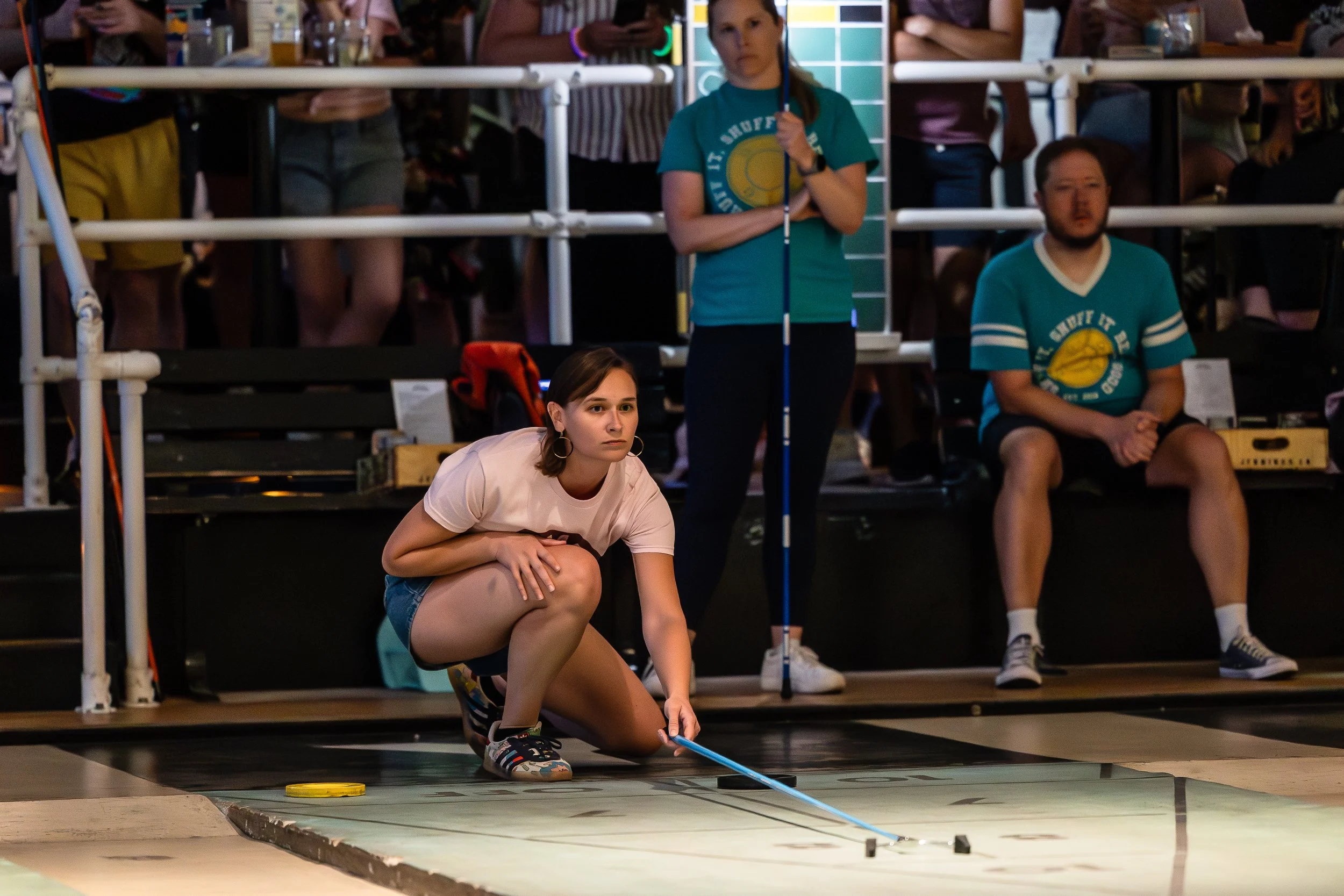 A young woman crouching on the floor using a guide stick to inspect a floor hockey rink, with a group of onlookers and officials watching behind her.