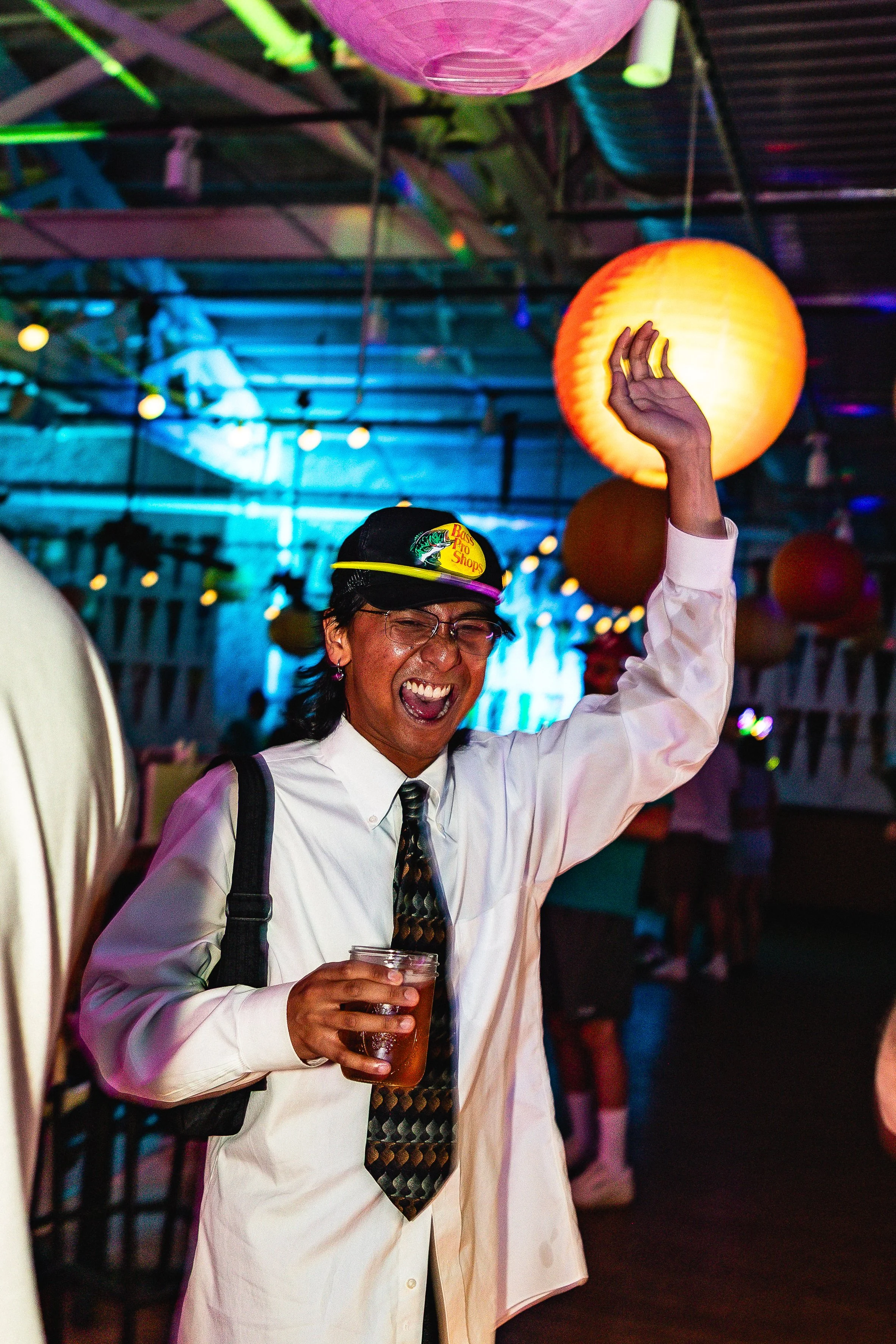 A man laughing and dancing at a party or celebration with colorful lanterns and string lights overhead. He is wearing glasses, a white shirt, a patterned tie, and a baseball cap, and holding a drink in his right hand with his left arm raised in the a