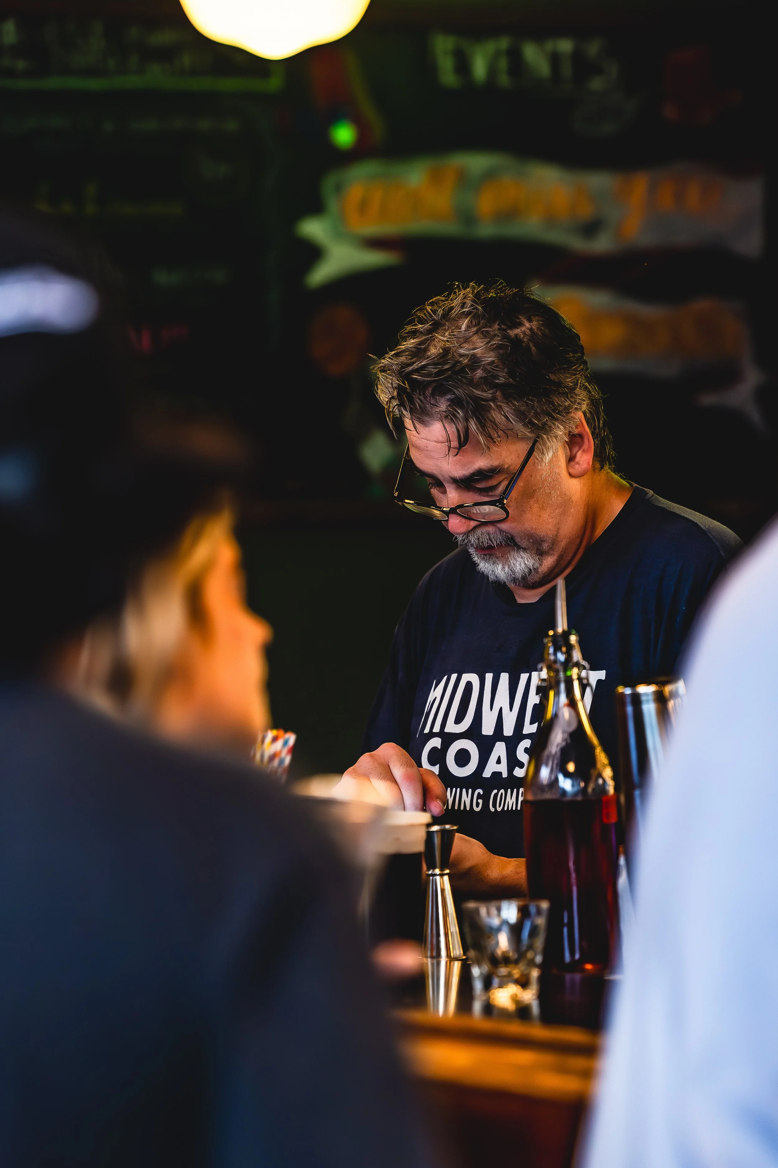 A bartender wearing glasses and a black T-shirt with 'MIDWEST COAST' written on it, preparing drinks in a bar with a dark background and colorful chalkboard menu.