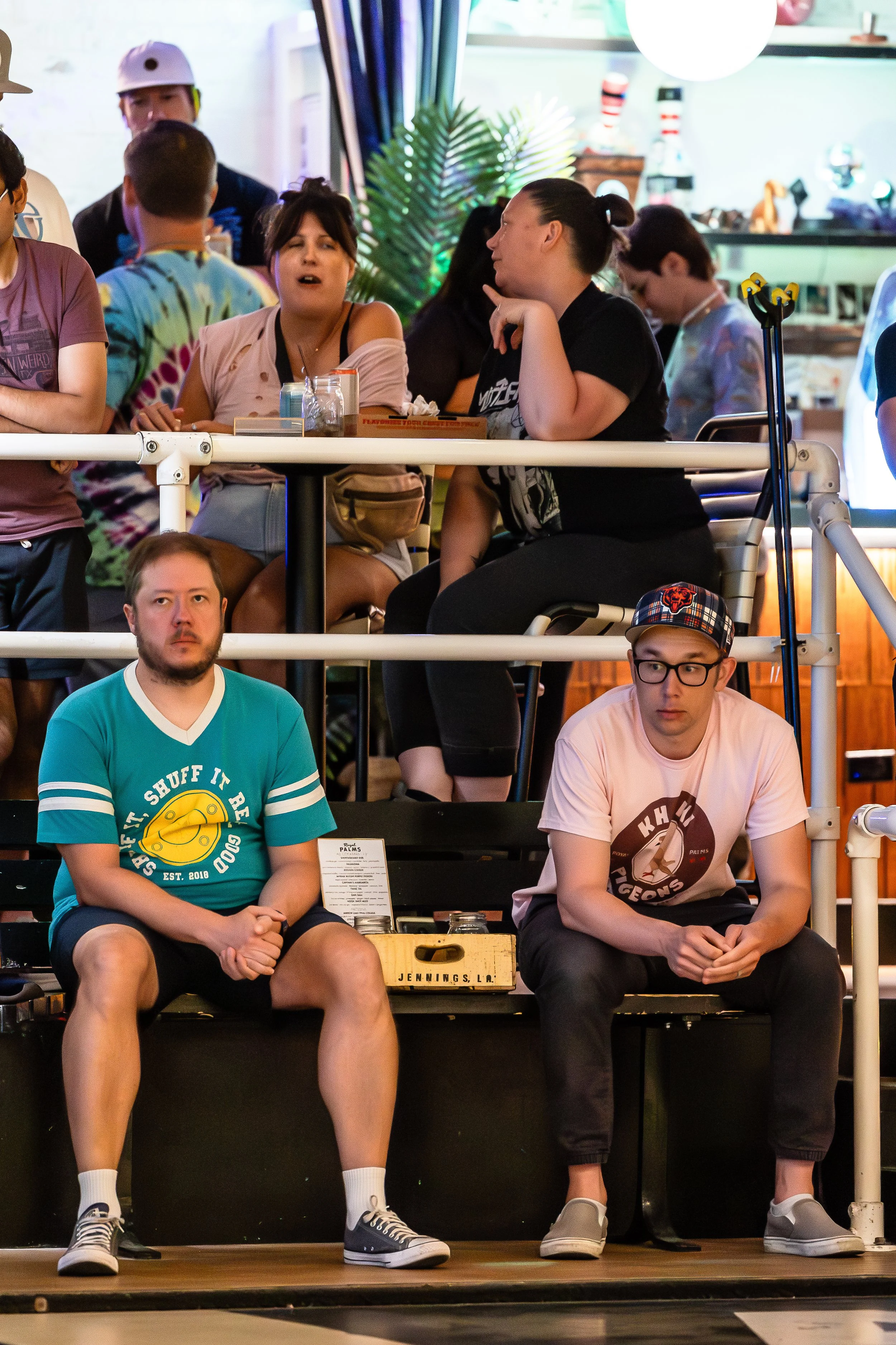 Group of people sitting in an indoor venue, some on a raised platform, watching an event. The background has colorful decor and lighting