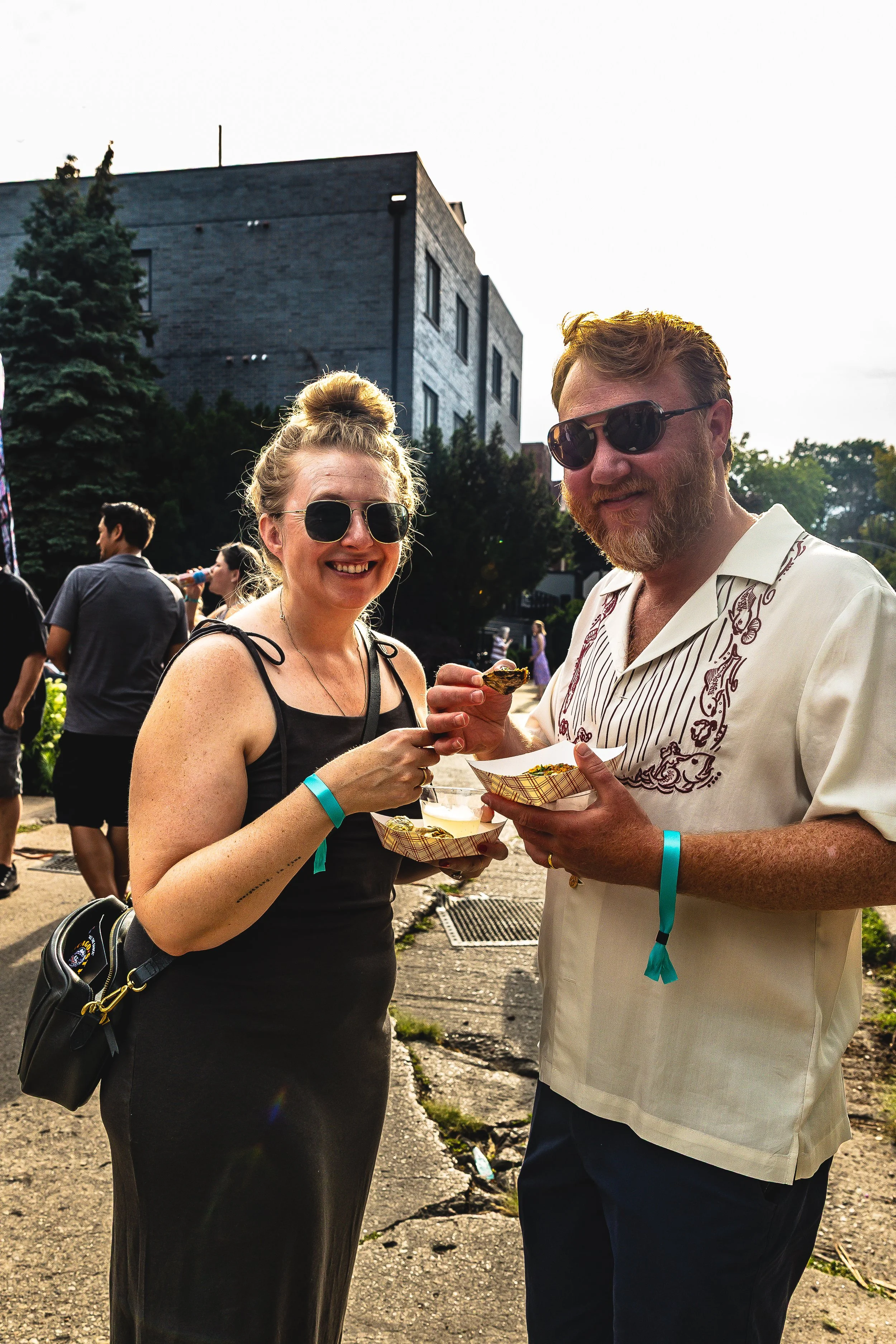 A smiling woman and man with sunglasses enjoying food at an outdoor event, holding food trays with a brick building and trees in the background.