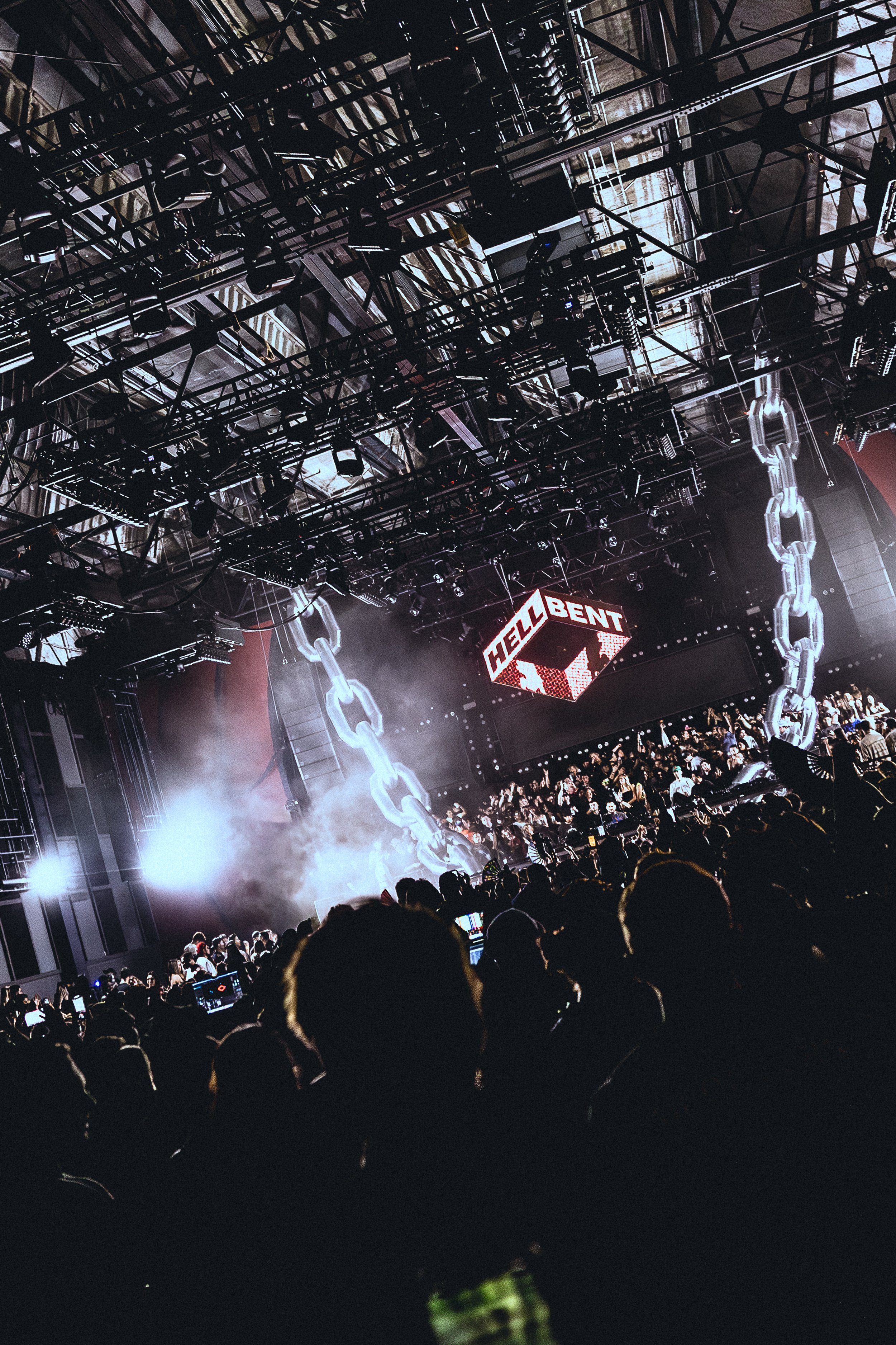 A concert scene with a large crowd, stage lighting, smoke effects, and a digital display with the words 'HELL BENT'. Large chains hang from the ceiling as part of the stage design.