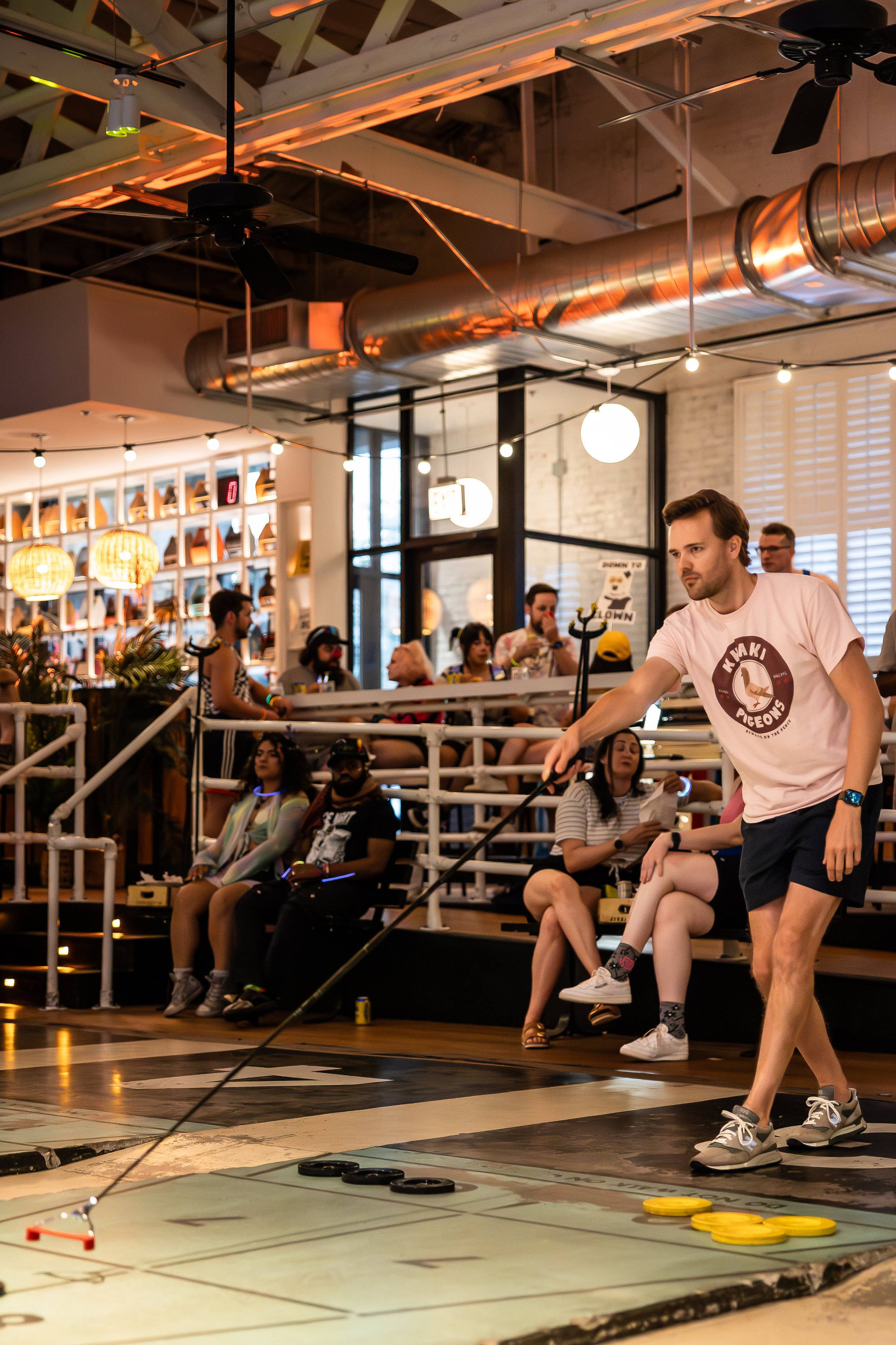A young man in a pink t-shirt and shorts playing shuffleboard on an indoor court while a group of people watch from the seating area. The scene is in a lively, modern space with industrial decor and warm lighting.