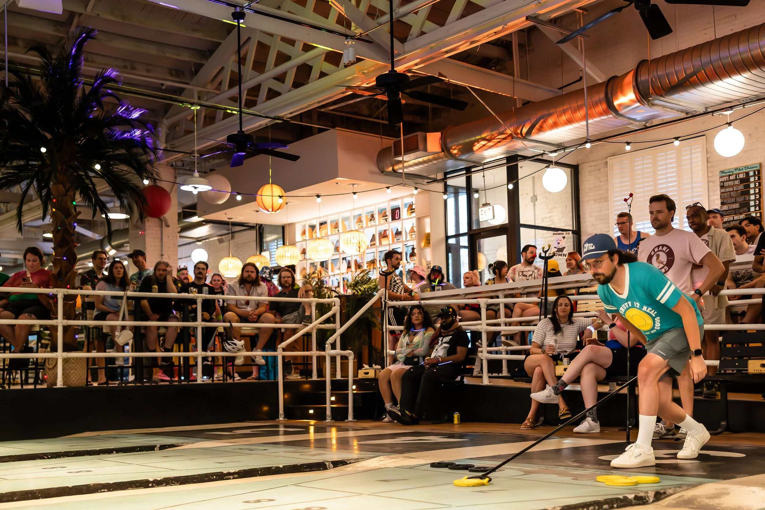 A man is playing shuffleboard in a lively, indoor social space with an audience watching. The setting has modern decor, warm lighting, and exposed ductwork on the ceiling.