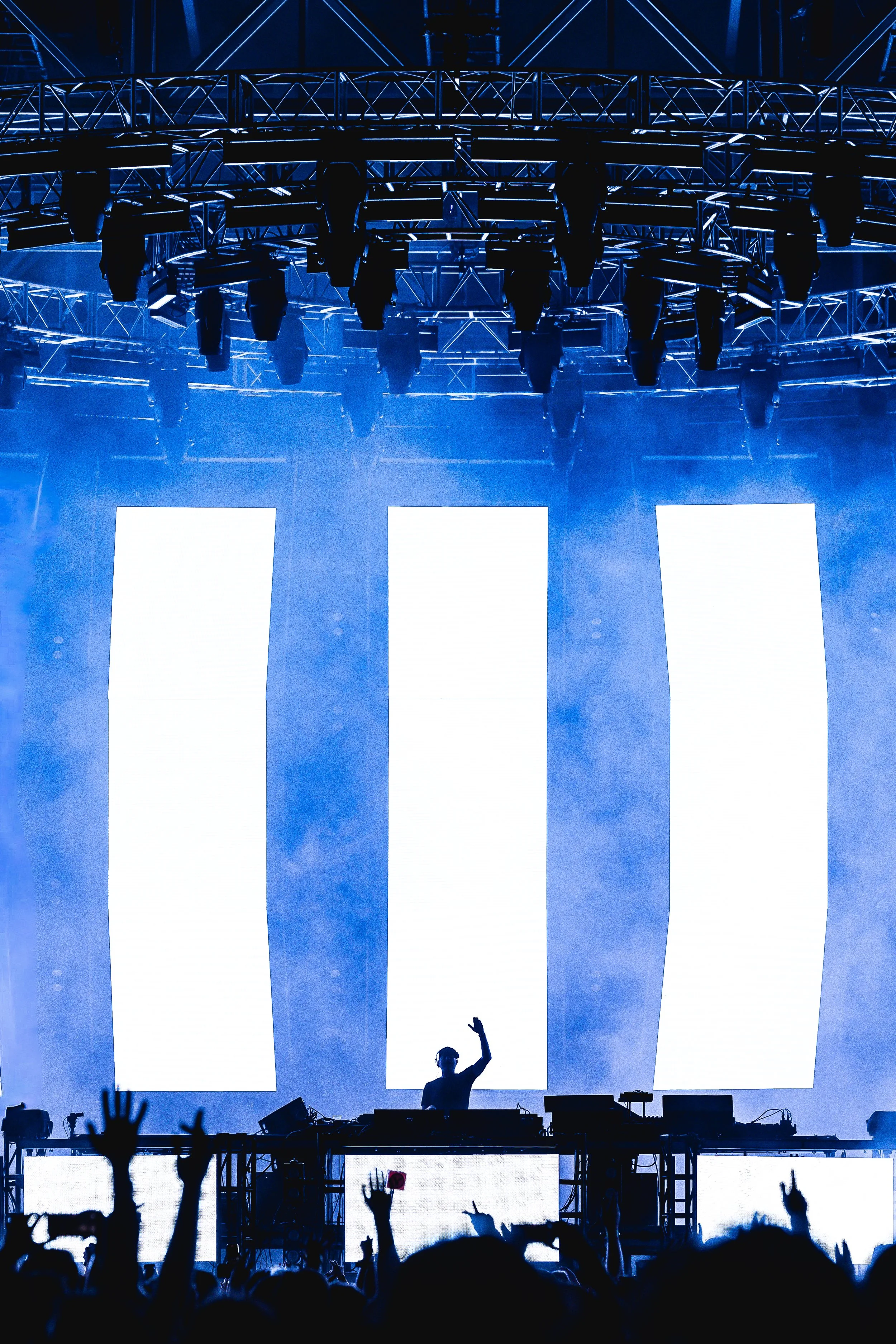 DJ performing on stage with bright LED screens, audience with raised hands, during a concert or music festival.