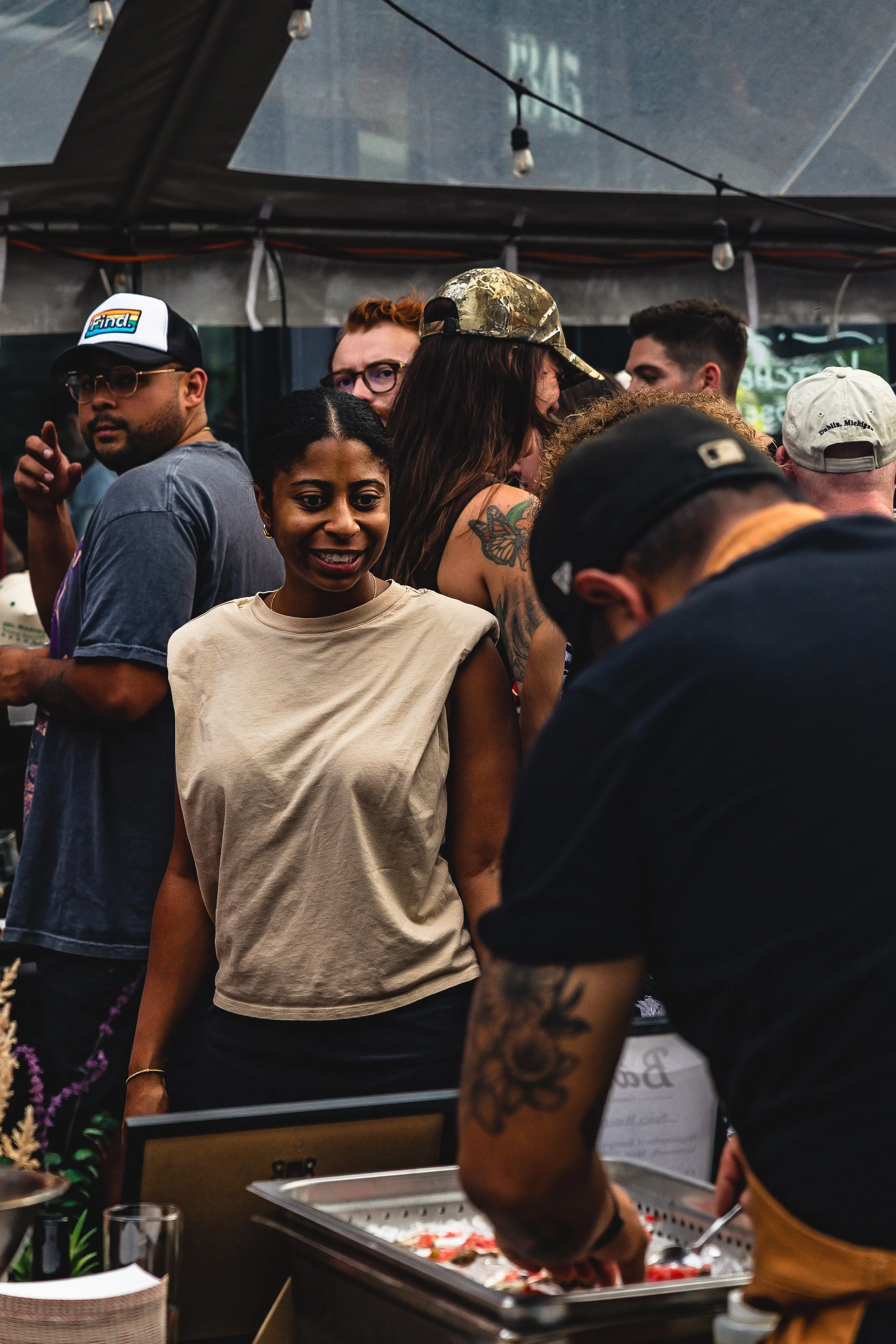 People at an outdoor event or market, some looking at a food or display table, with diverse individuals including a woman with a butterfly tattoo and a man with tattoos on his arm, under a large tent.