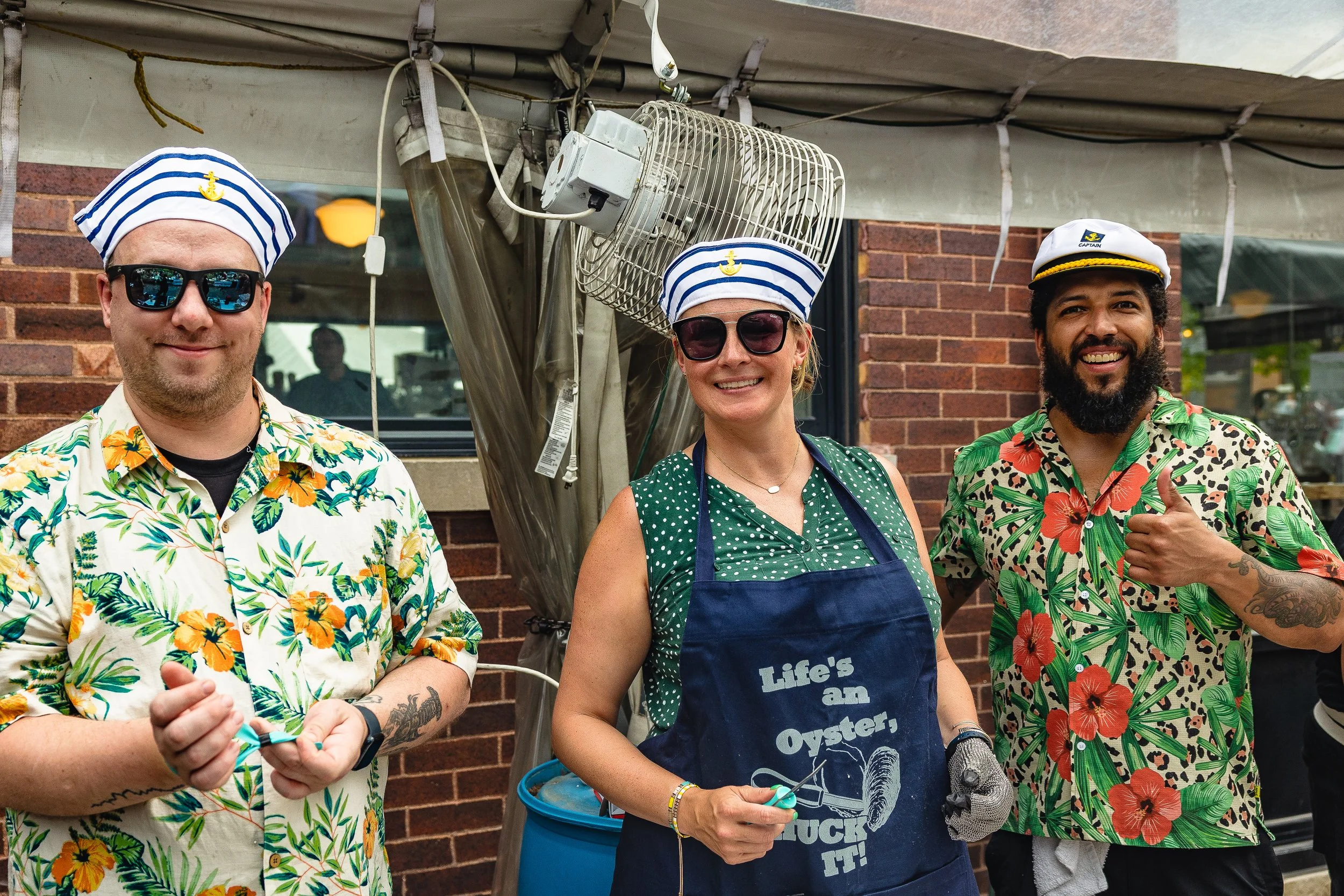 Three people dressed in Hawaiian shirts and sailor hats, standing outdoors in front of a brick wall, smiling and posing for the photo.