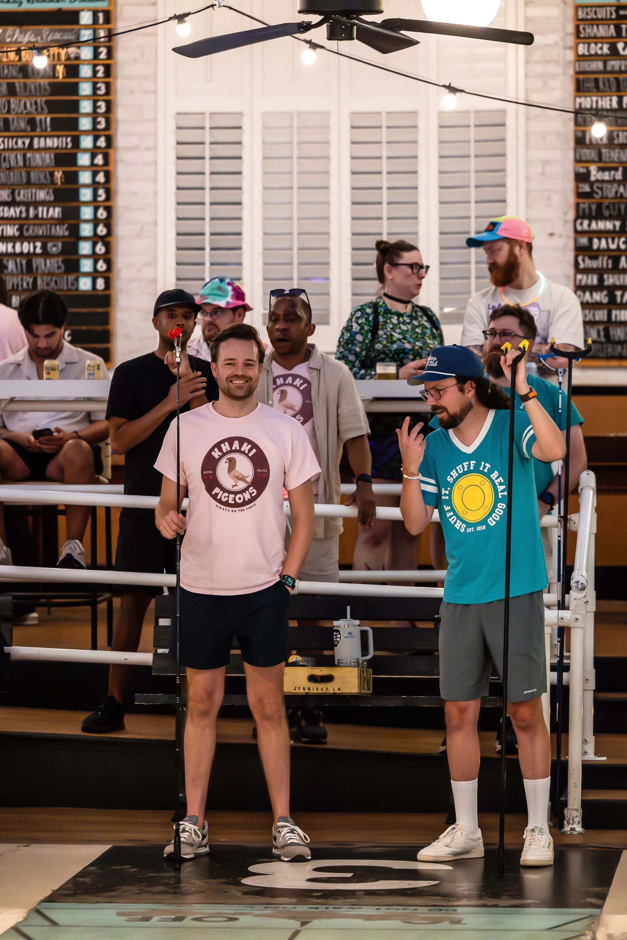 A group of people at a bowling alley. Two men in the foreground are holding bowling balls and wearing casual shirts with slogans, smiling, and talking. Several others are in the background near the scorer's table, with some seated and using their pho