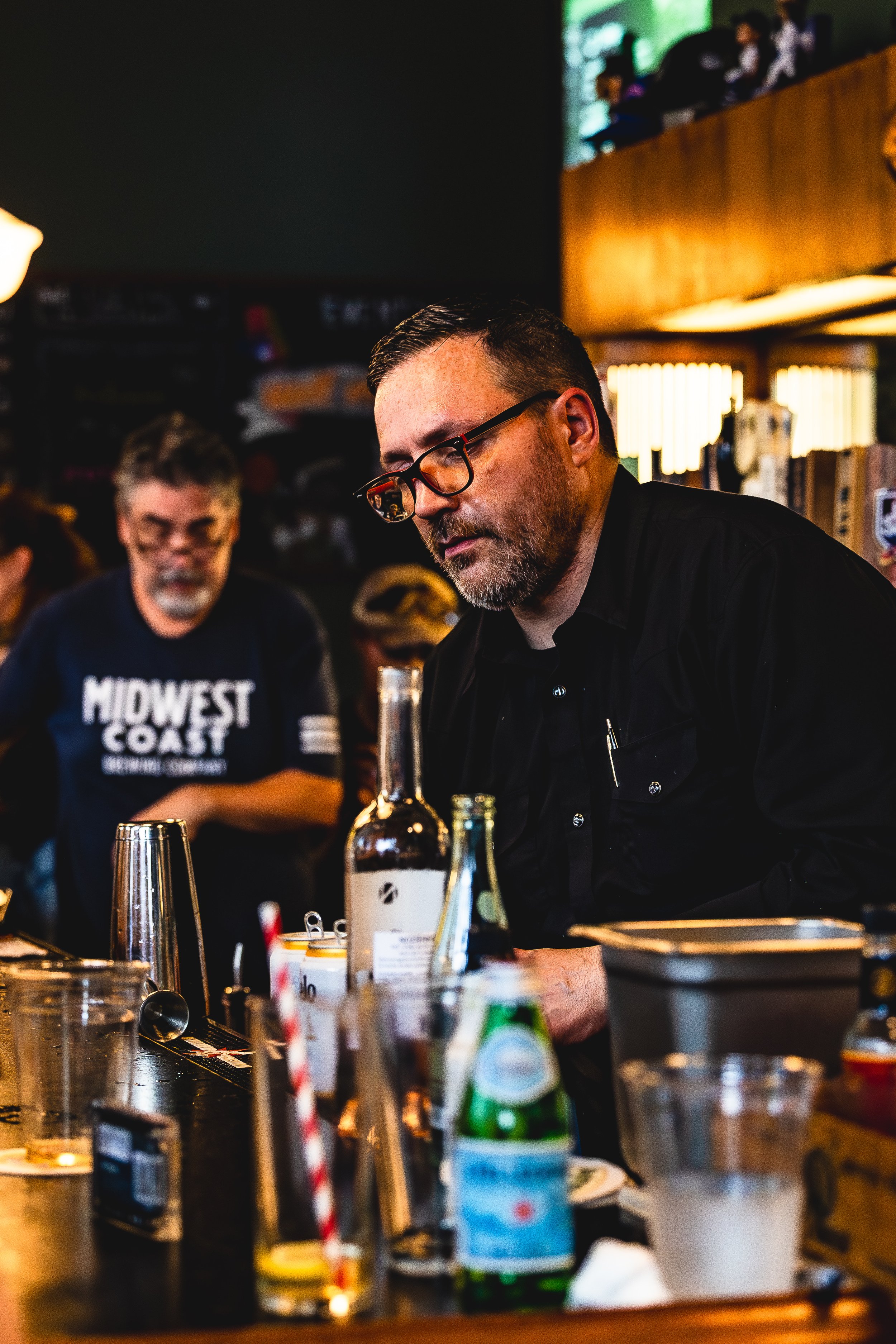 A man with glasses and a beard working behind a bar counter, surrounded by bottles and glasses, in a dimly lit bar or restaurant with other people in the background.